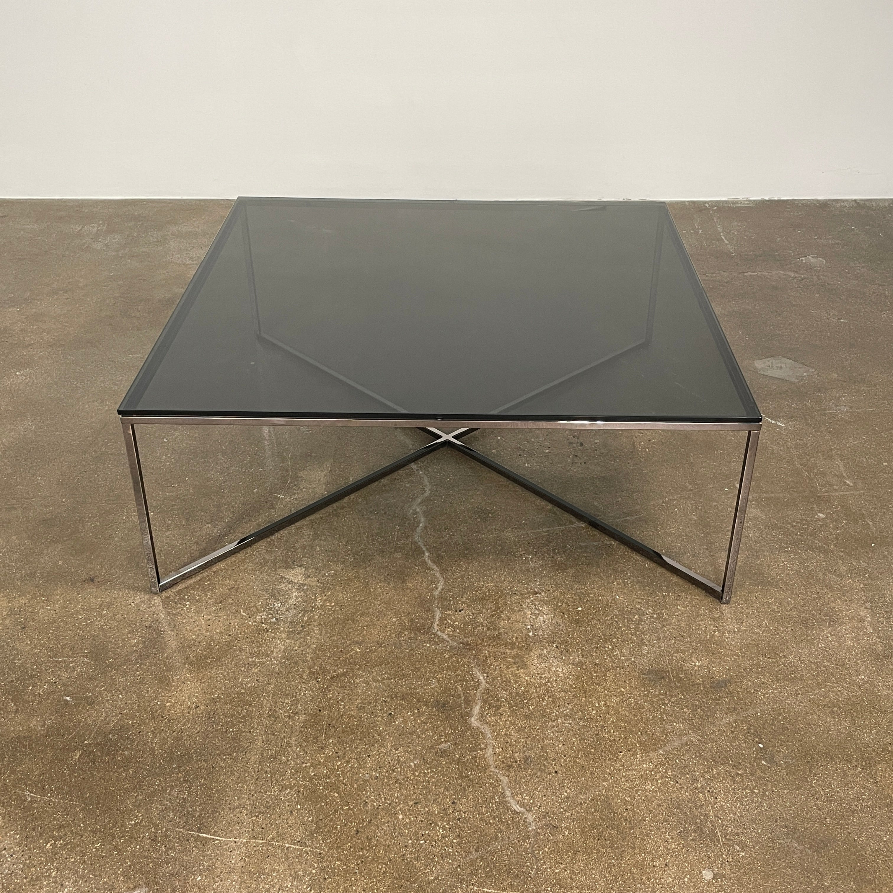 The Smoked Glass Coffee Table features a square design with a contemporary metal frame and crossed support bars, shown against a plain white background. Brand: Unknown.