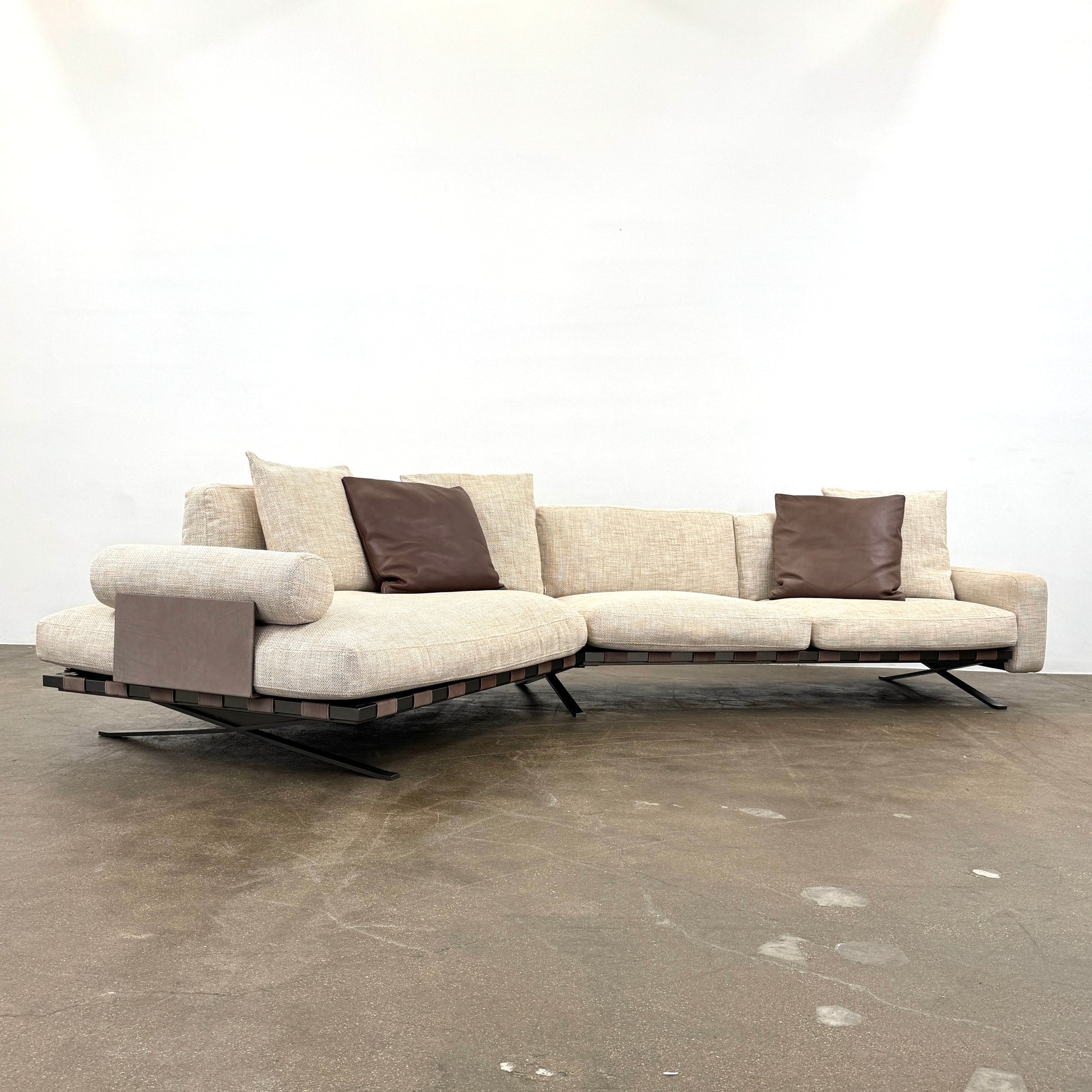 The Driade Fenix sofa by Driade is a beige L-shaped modular design with three seat cushions, black legs, and four throw pillows—two brown, two beige—highlighting its architectural lightness against a plain white backdrop.