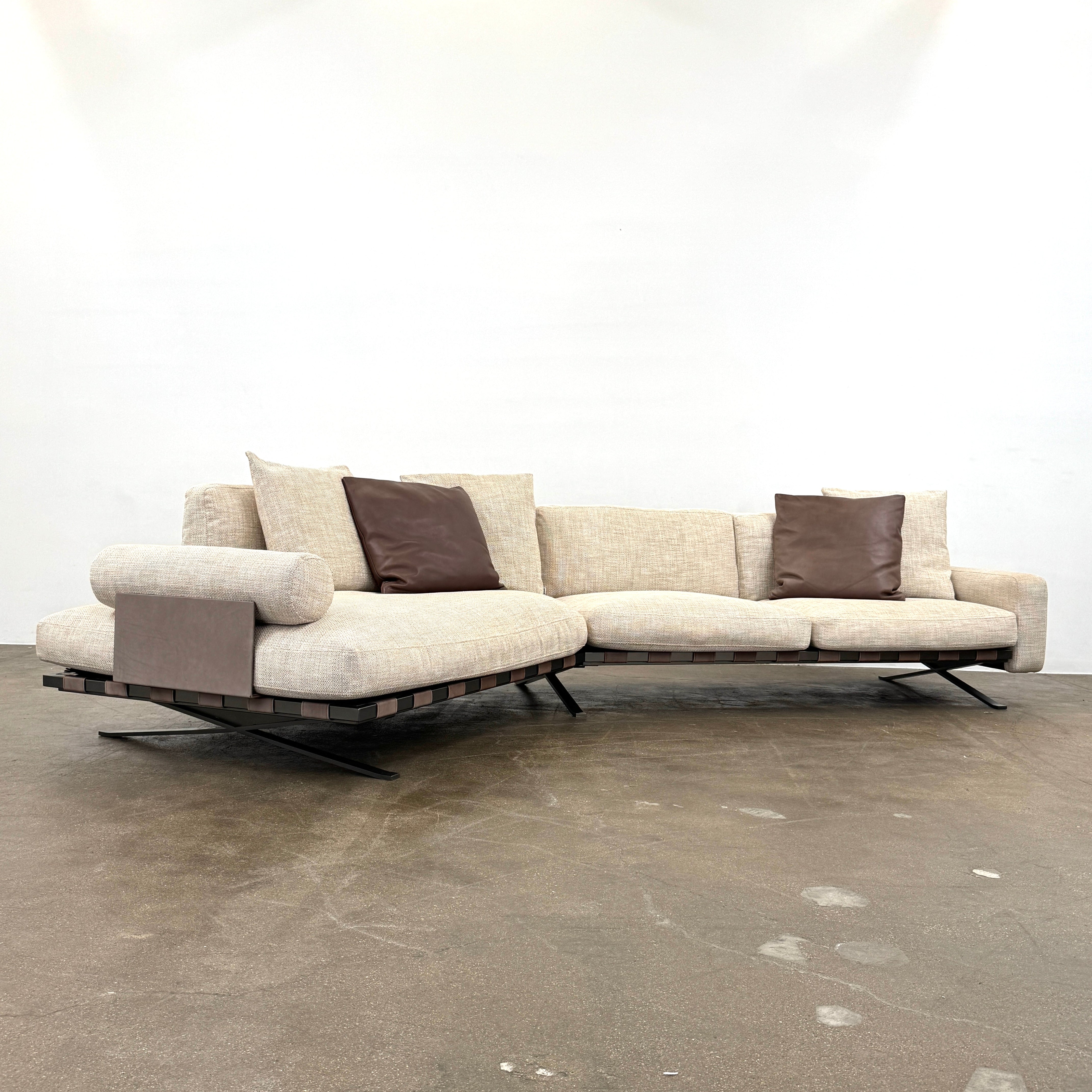 The Driade Fenix sofa by Driade is a beige L-shaped modular design with three seat cushions, black legs, and four throw pillows—two brown, two beige—highlighting its architectural lightness against a plain white backdrop.