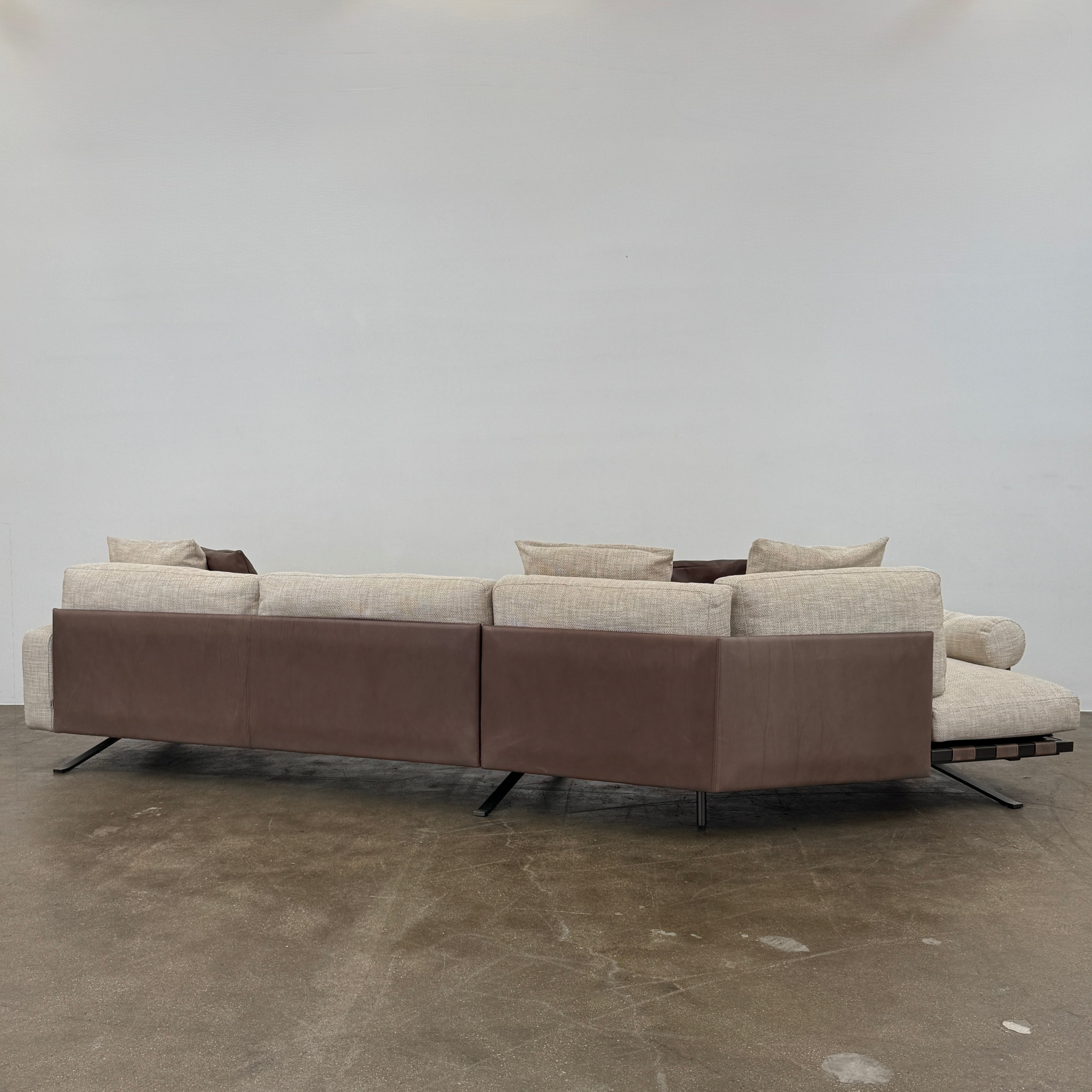 The Driade Fenix sofa by Driade is a beige L-shaped modular design with three seat cushions, black legs, and four throw pillows—two brown, two beige—highlighting its architectural lightness against a plain white backdrop.