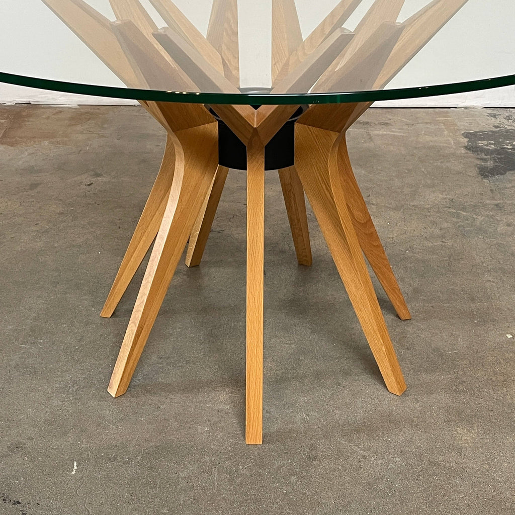 A Roche Bobois Roche Bobois Aster dining table features a round glass tabletop supported by a wooden base with several spokes radiating outwards, designed by French-Algerian designer Reda Amalou.