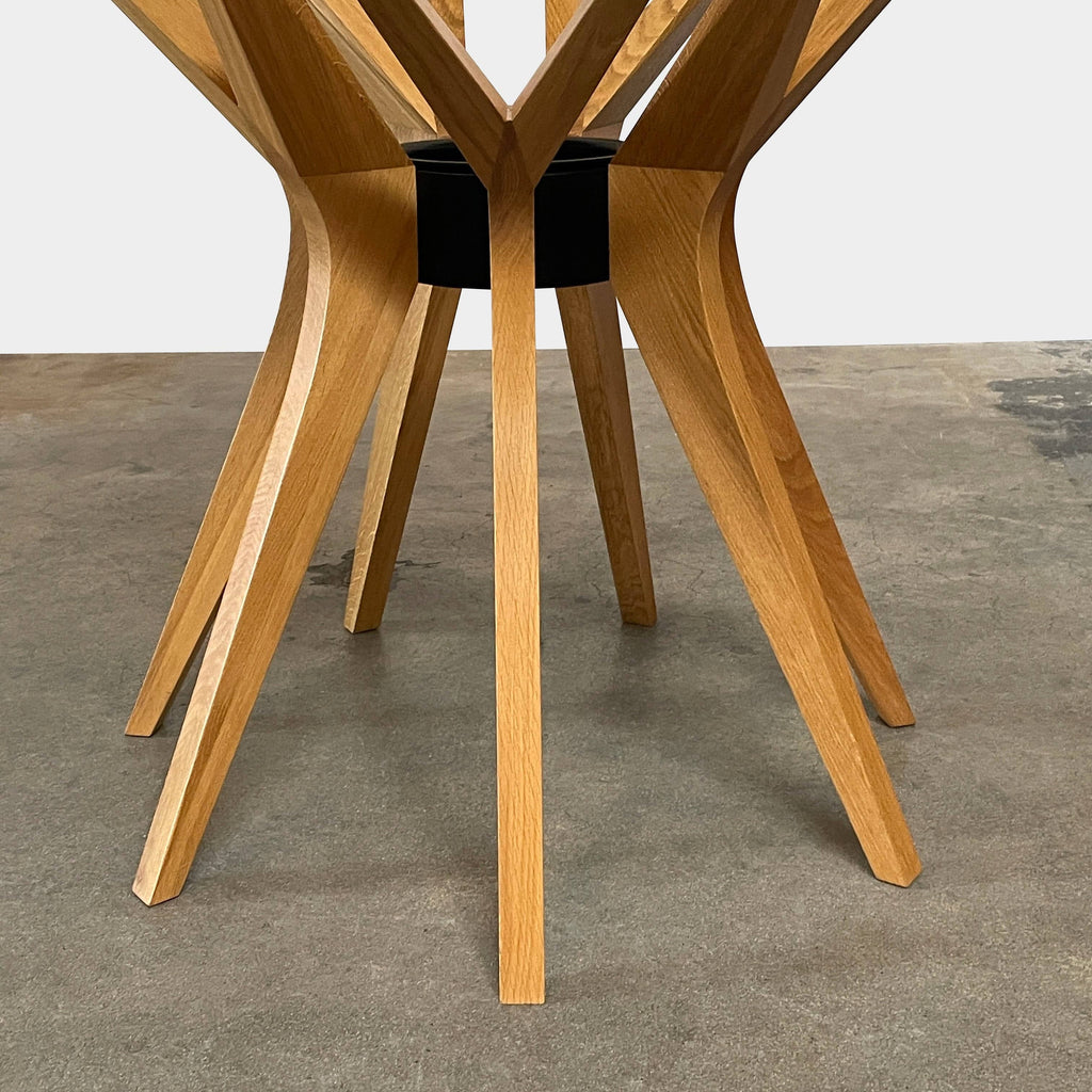 A Roche Bobois Roche Bobois Aster dining table features a round glass tabletop supported by a wooden base with several spokes radiating outwards, designed by French-Algerian designer Reda Amalou.