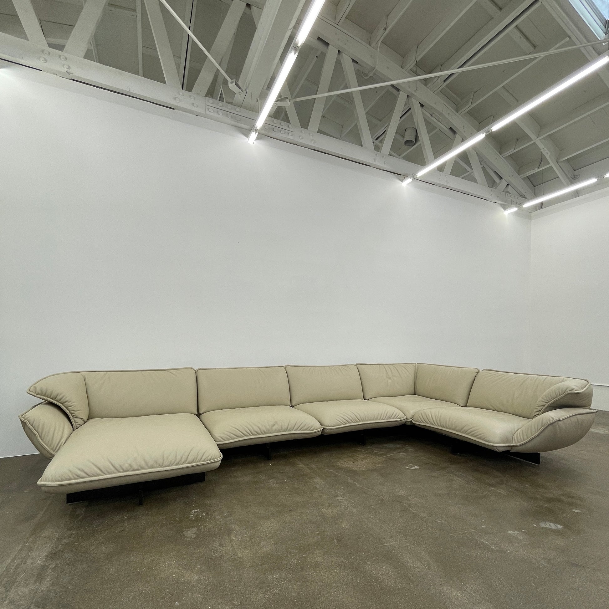 The Cassina Superbeam 5 Piece Leather Sectional in beige, featuring plush cushions, is arranged in a spacious room with white walls, exposed Superbeam ceiling, and overhead lighting.