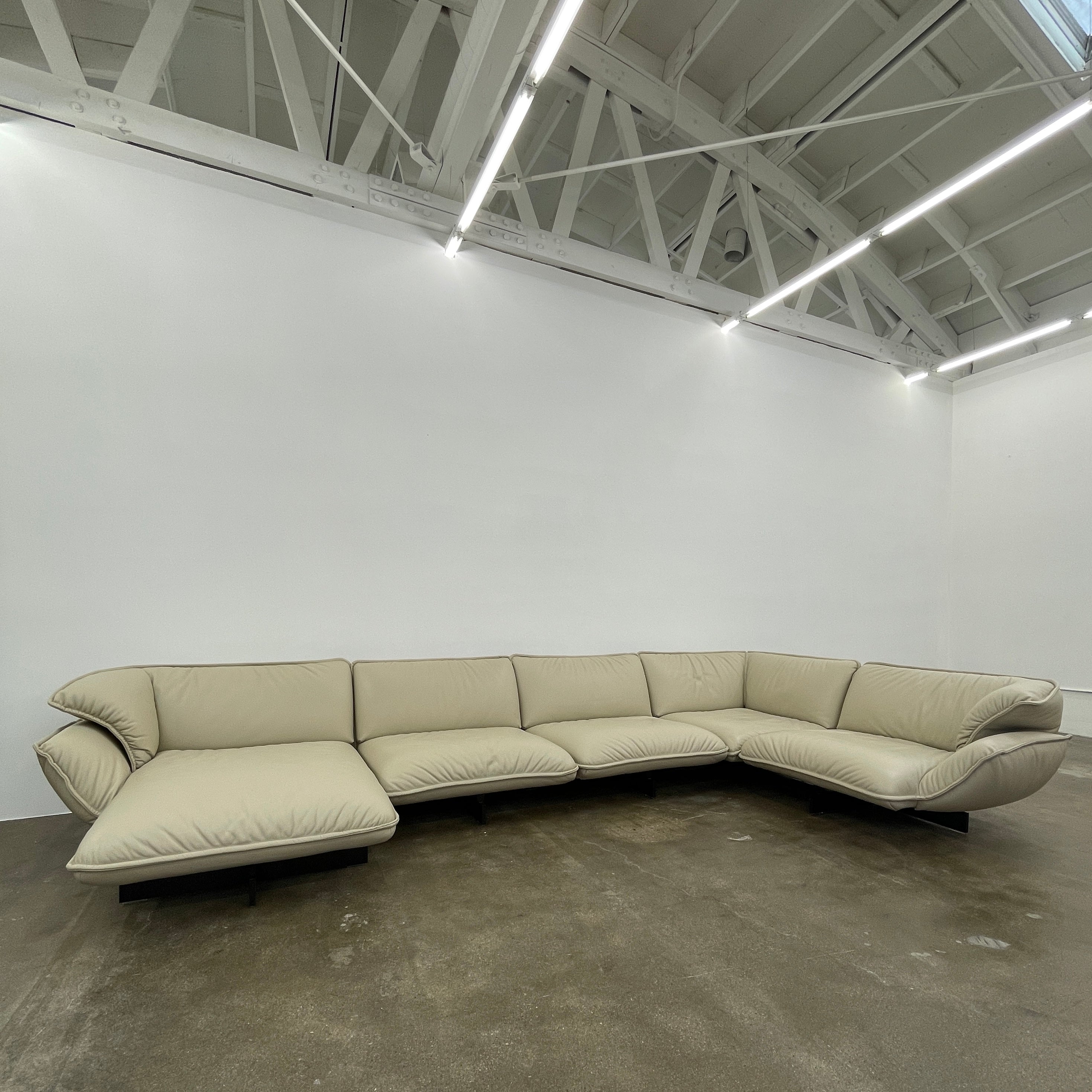 The Cassina Superbeam 5 Piece Leather Sectional in beige, featuring plush cushions, is arranged in a spacious room with white walls, exposed Superbeam ceiling, and overhead lighting.