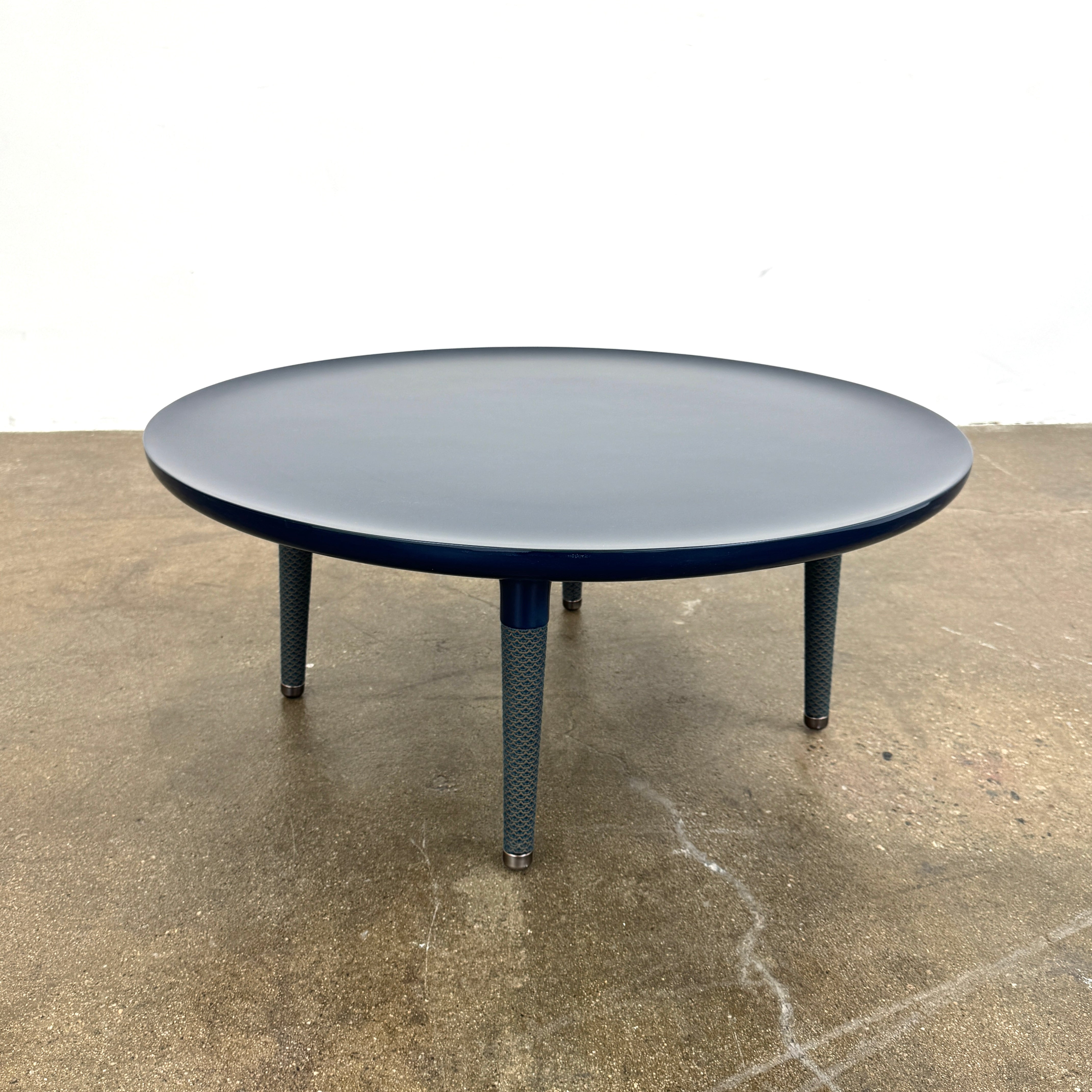 The Roche Bobois Globe Trotter La Parisienne cocktail table features a round, dark blue top and three textured legs, shown against a light gray background.