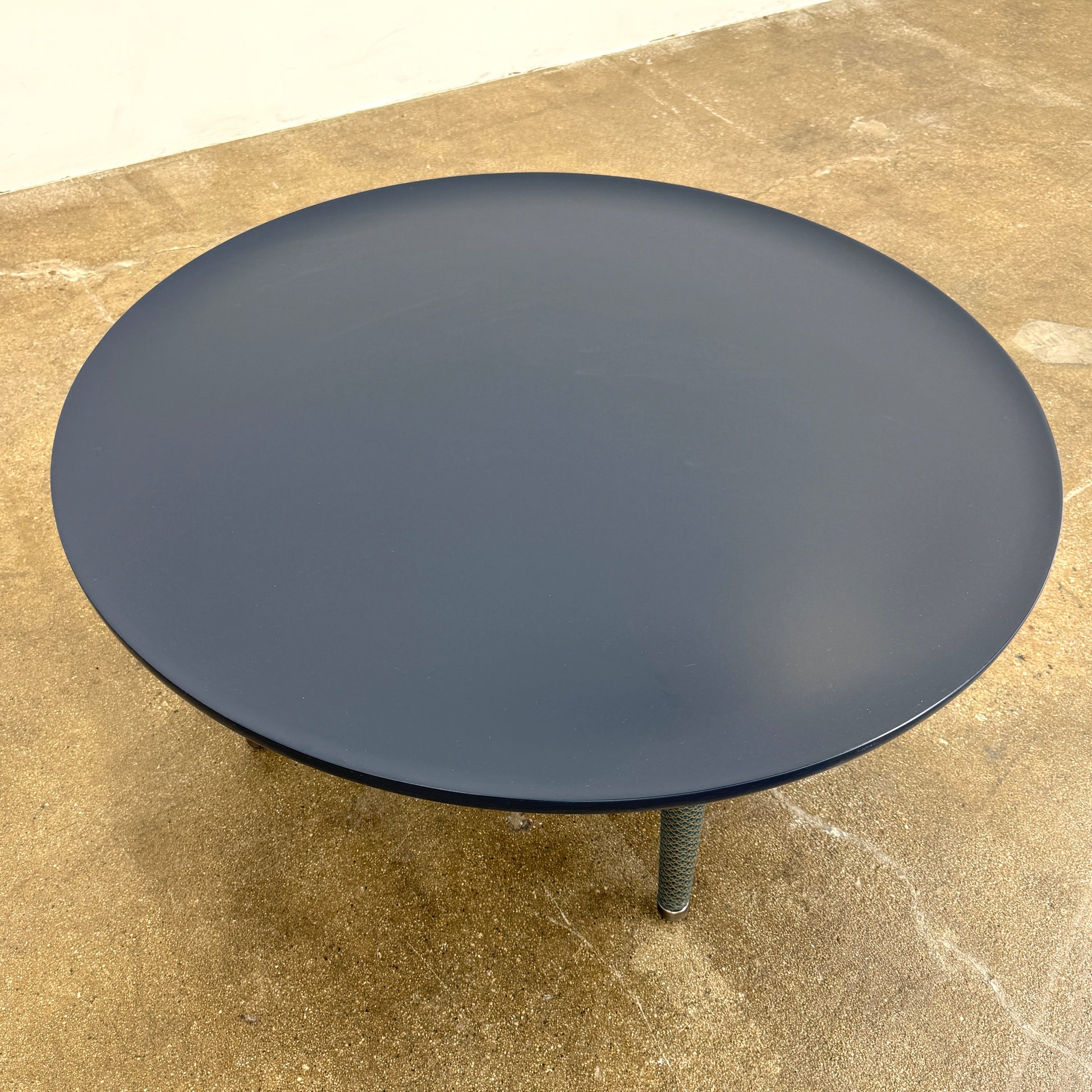 The Roche Bobois Globe Trotter La Parisienne cocktail table features a round, dark blue top and three textured legs, shown against a light gray background.