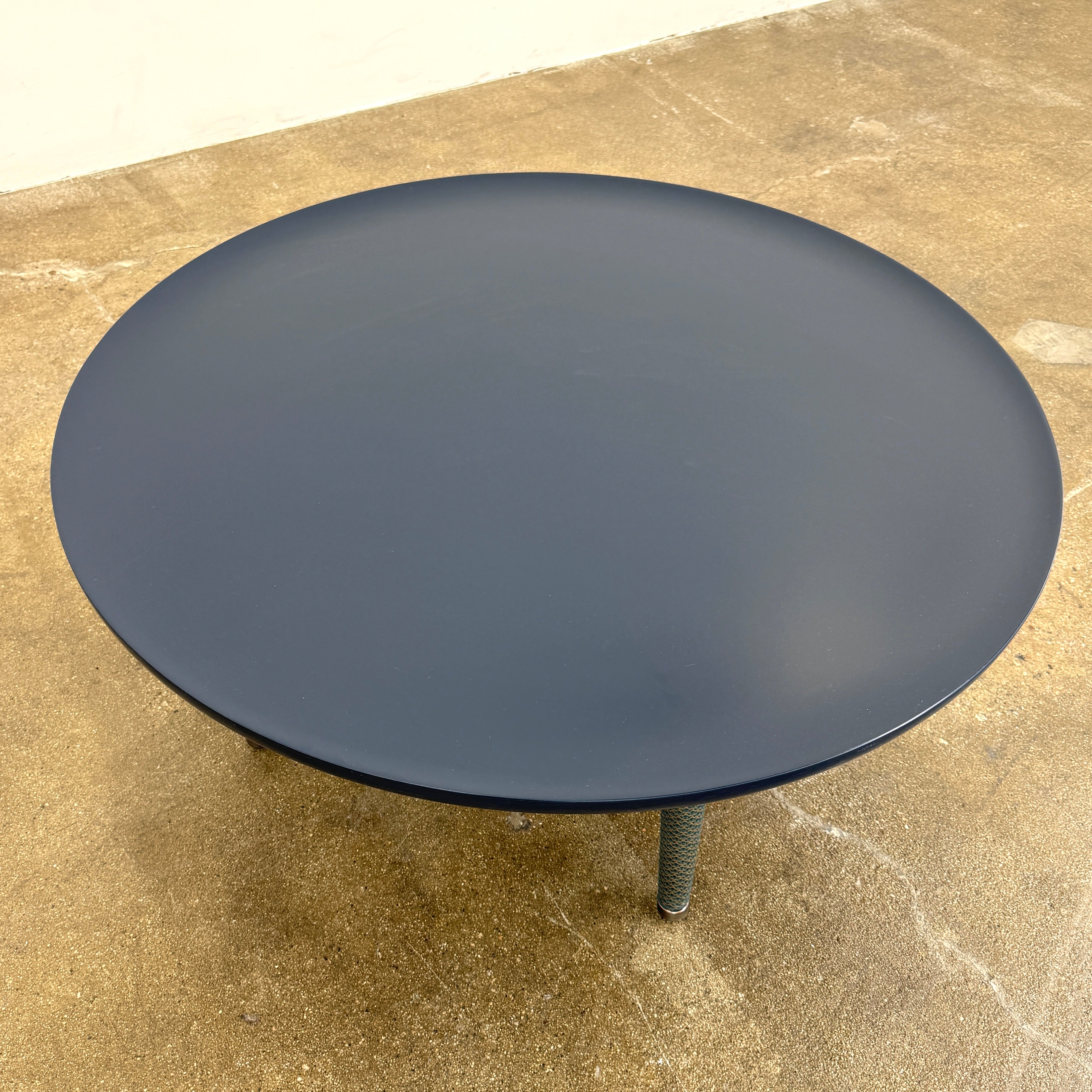 The Roche Bobois Globe Trotter La Parisienne cocktail table features a round, dark blue top and three textured legs, shown against a light gray background.