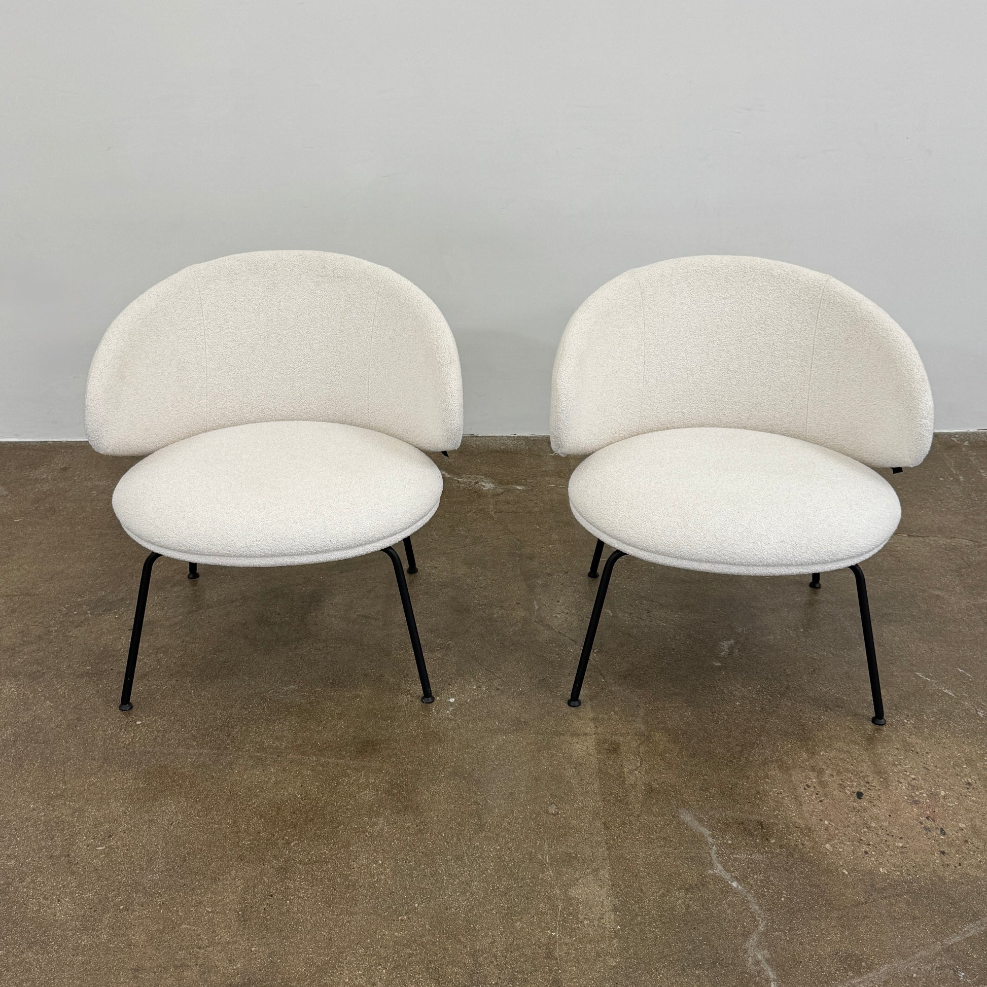 Two Arrmet Belle Lounge chairs by Arrmet, featuring white cushions, rounded backs, and black metal legs, are placed side by side on a brown concrete floor against a plain light gray wall.