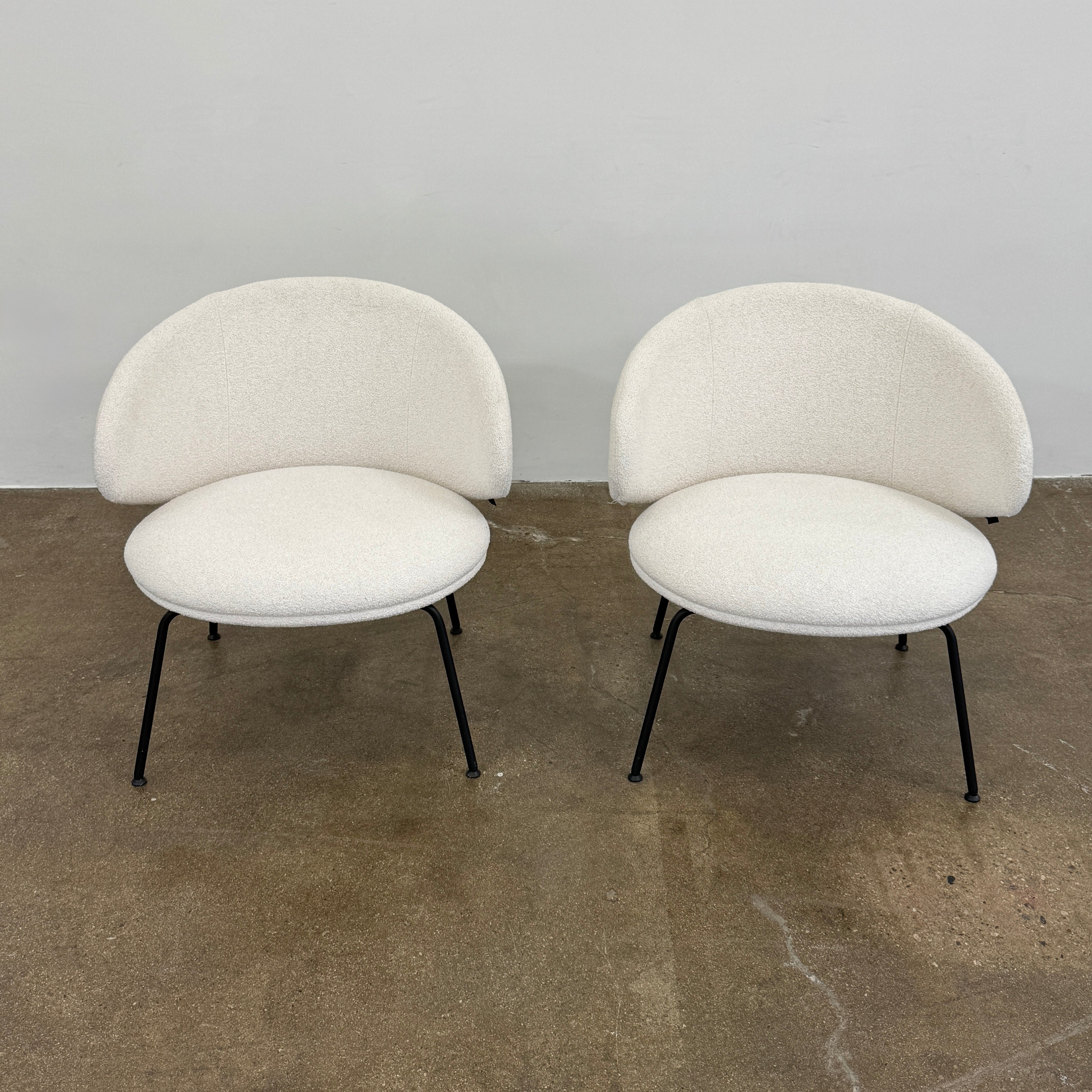 Two Arrmet Belle Lounge chairs by Arrmet, featuring white cushions, rounded backs, and black metal legs, are placed side by side on a brown concrete floor against a plain light gray wall.