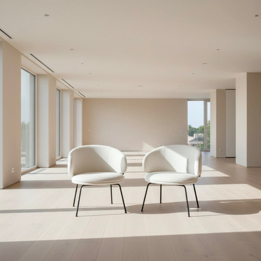 Two Arrmet Belle Lounge chairs by Arrmet, featuring white cushions, rounded backs, and black metal legs, are placed side by side on a brown concrete floor against a plain light gray wall.