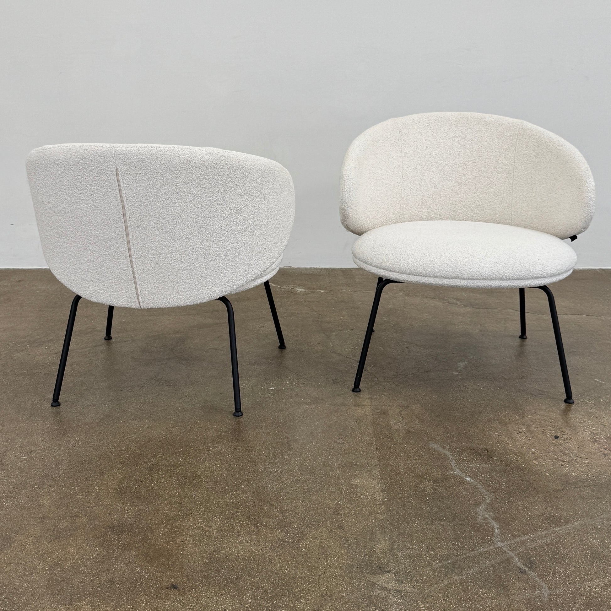 Two Arrmet Belle Lounge chairs by Arrmet, featuring white cushions, rounded backs, and black metal legs, are placed side by side on a brown concrete floor against a plain light gray wall.