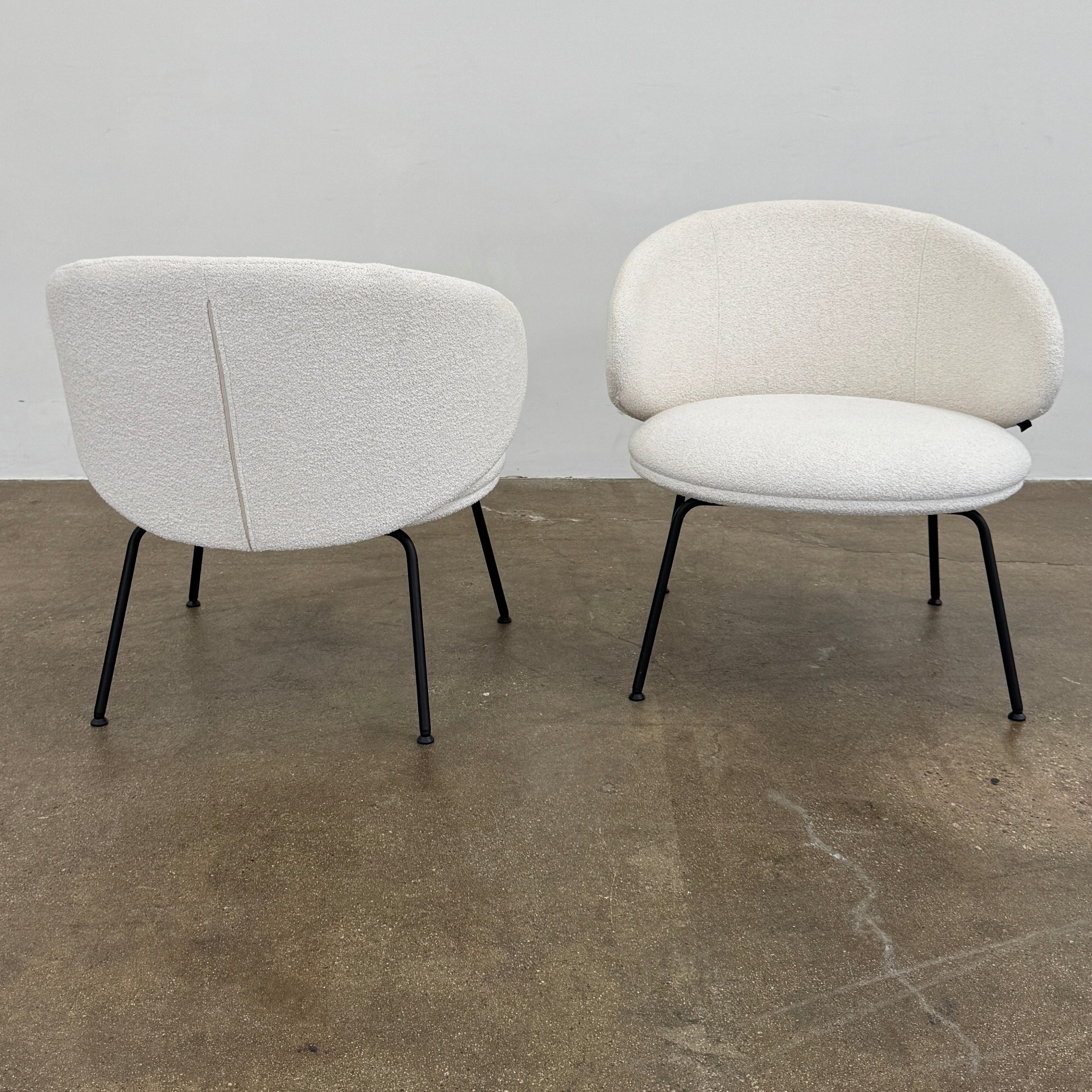 Two Arrmet Belle Lounge chairs by Arrmet, featuring white cushions, rounded backs, and black metal legs, are placed side by side on a brown concrete floor against a plain light gray wall.