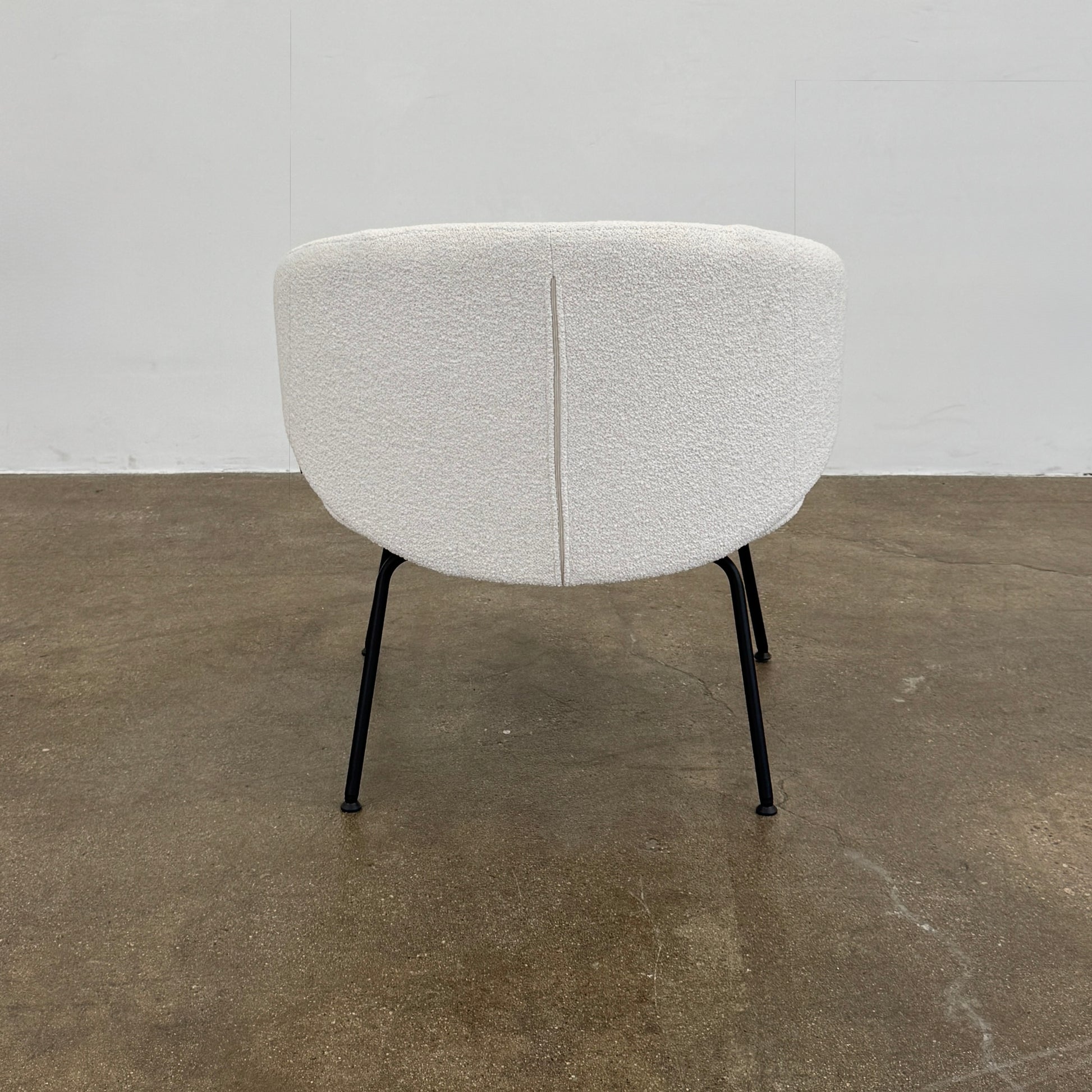 Two Arrmet Belle Lounge chairs by Arrmet, featuring white cushions, rounded backs, and black metal legs, are placed side by side on a brown concrete floor against a plain light gray wall.