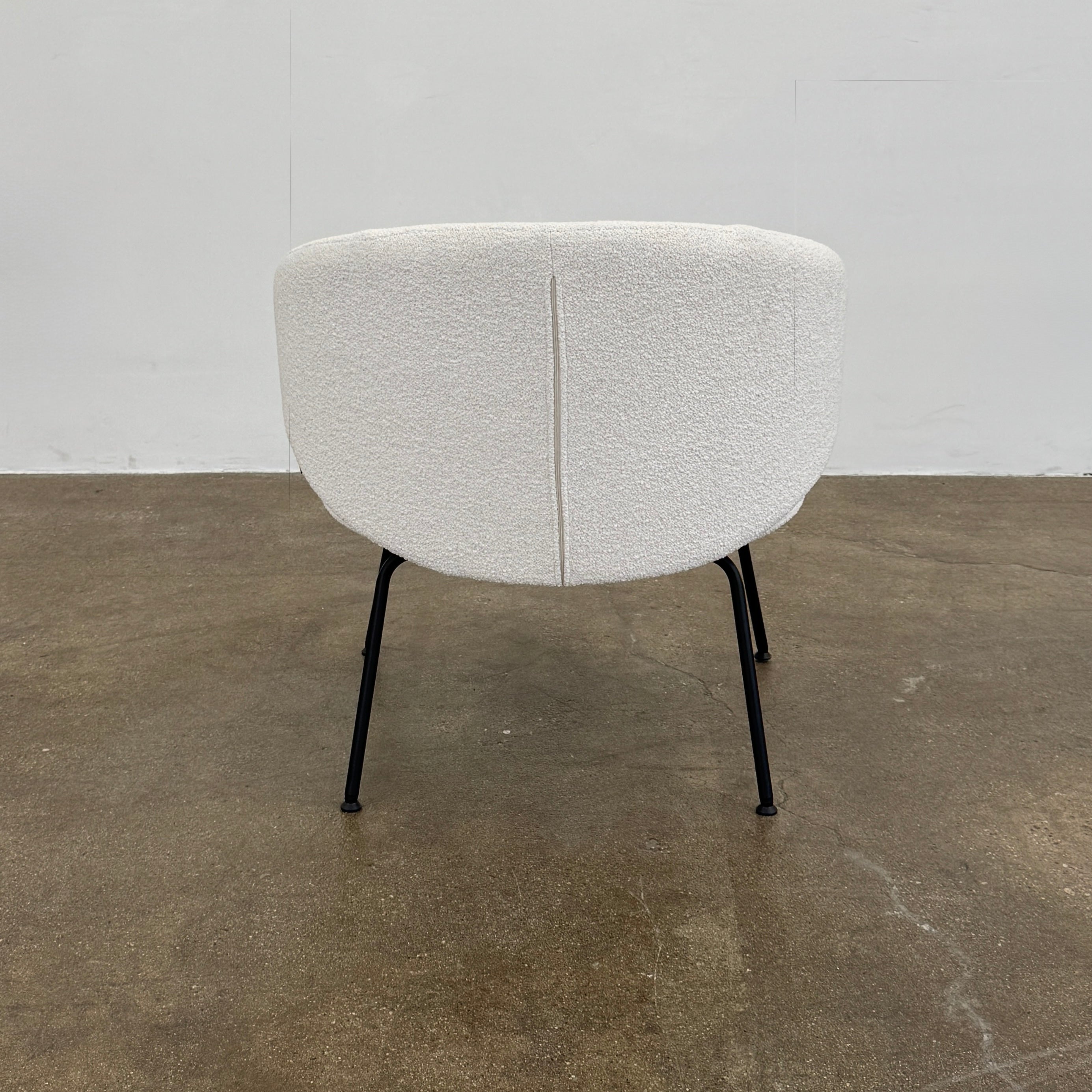 Two Arrmet Belle Lounge chairs by Arrmet, featuring white cushions, rounded backs, and black metal legs, are placed side by side on a brown concrete floor against a plain light gray wall.