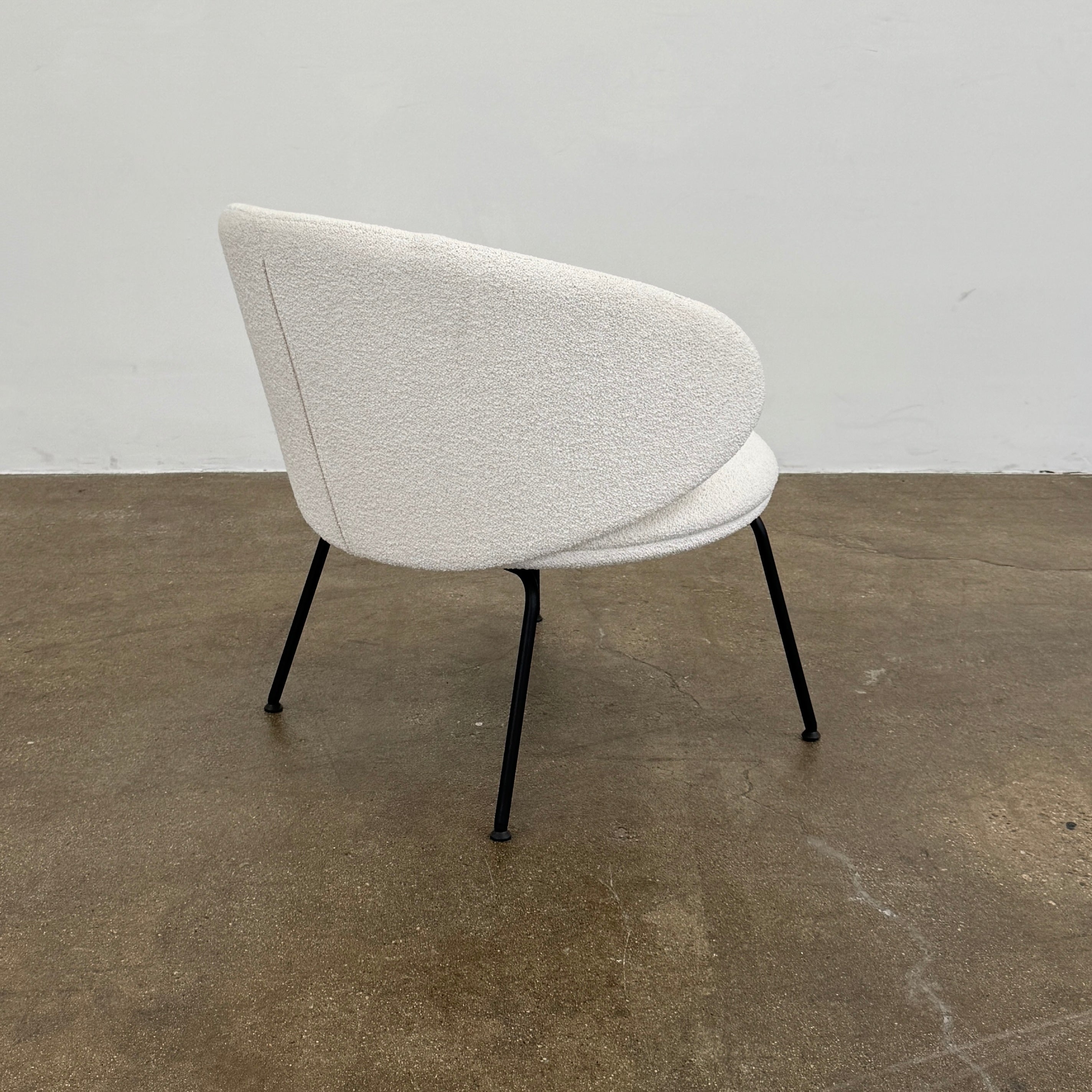 Two Arrmet Belle Lounge chairs by Arrmet, featuring white cushions, rounded backs, and black metal legs, are placed side by side on a brown concrete floor against a plain light gray wall.