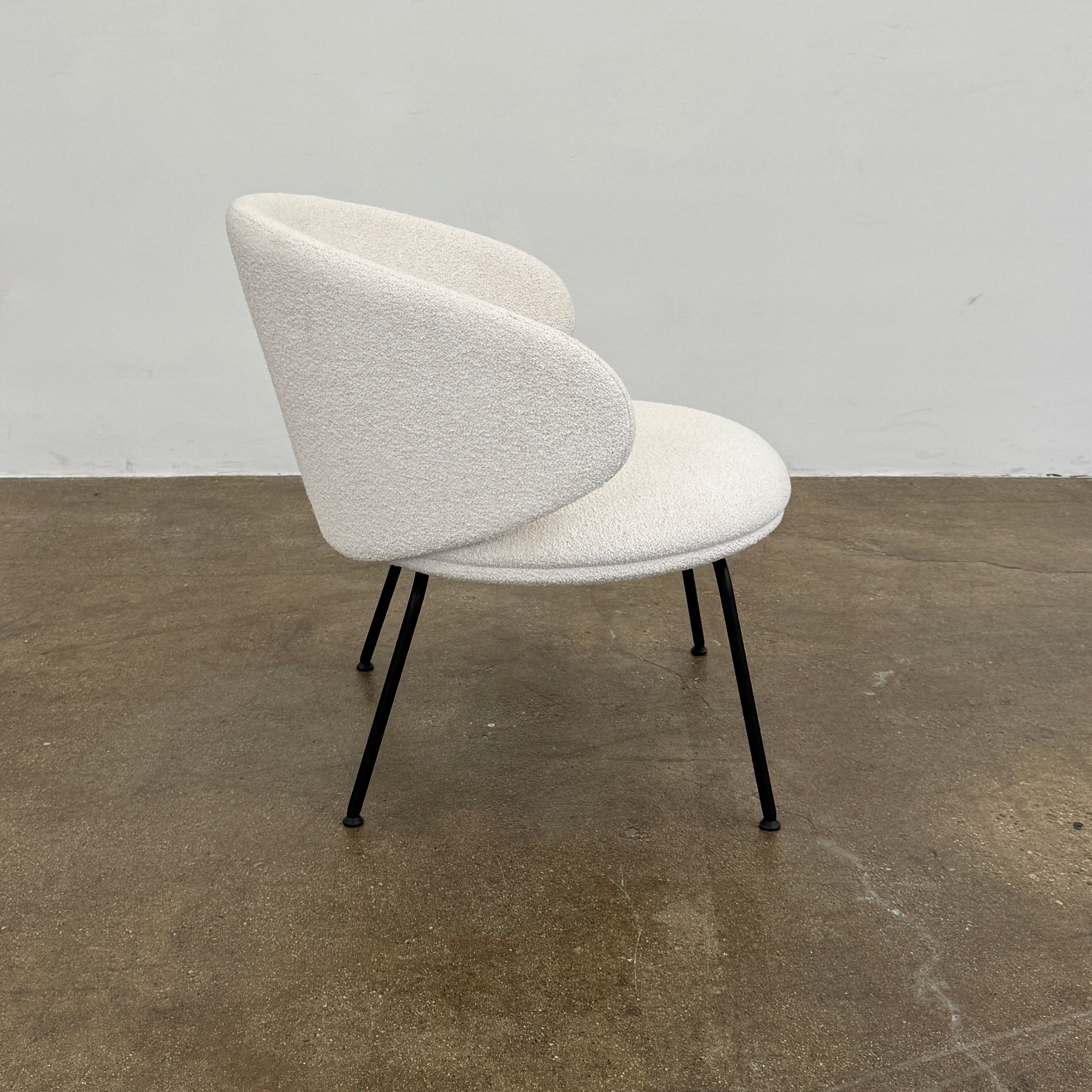 Two Arrmet Belle Lounge chairs by Arrmet, featuring white cushions, rounded backs, and black metal legs, are placed side by side on a brown concrete floor against a plain light gray wall.