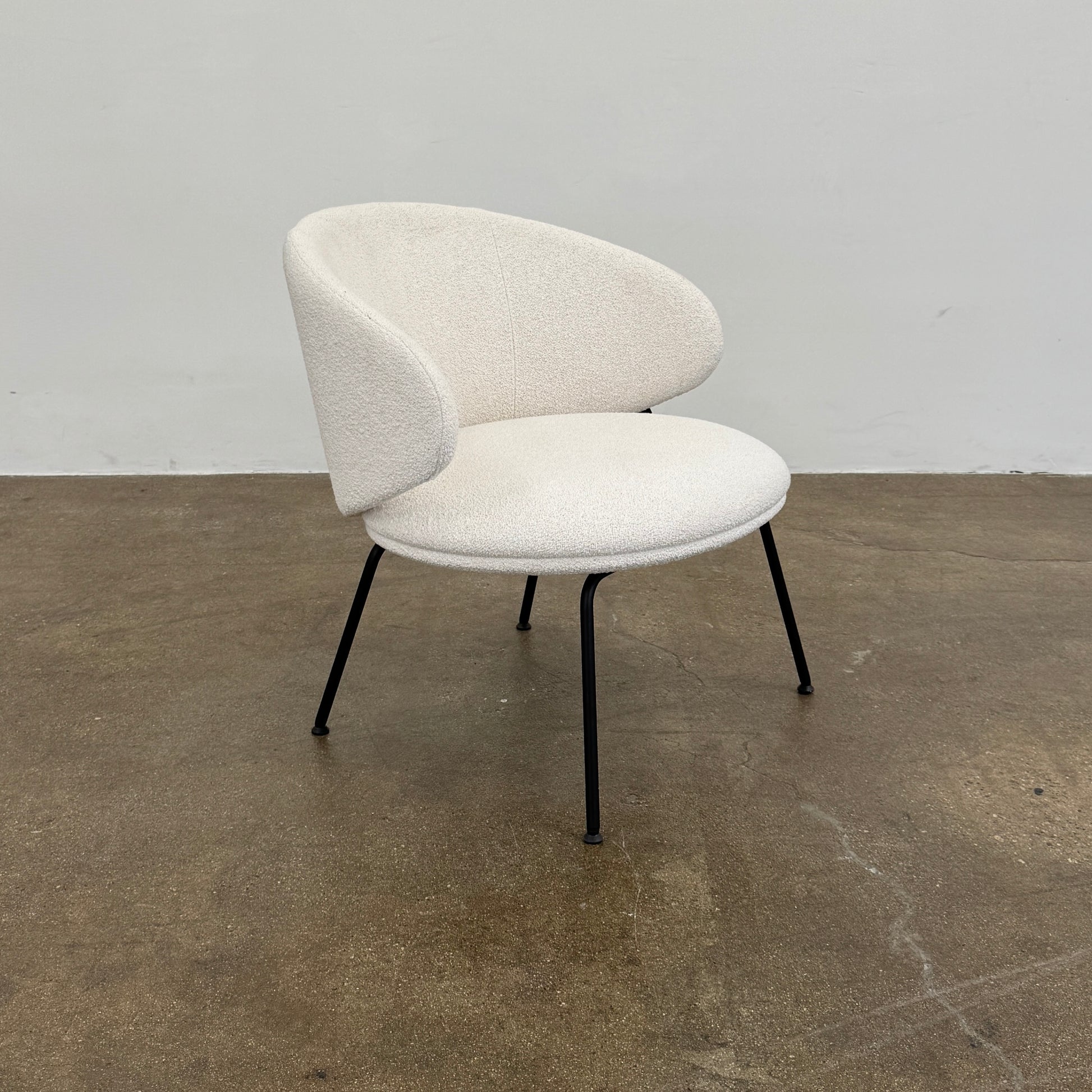 Two Arrmet Belle Lounge chairs by Arrmet, featuring white cushions, rounded backs, and black metal legs, are placed side by side on a brown concrete floor against a plain light gray wall.