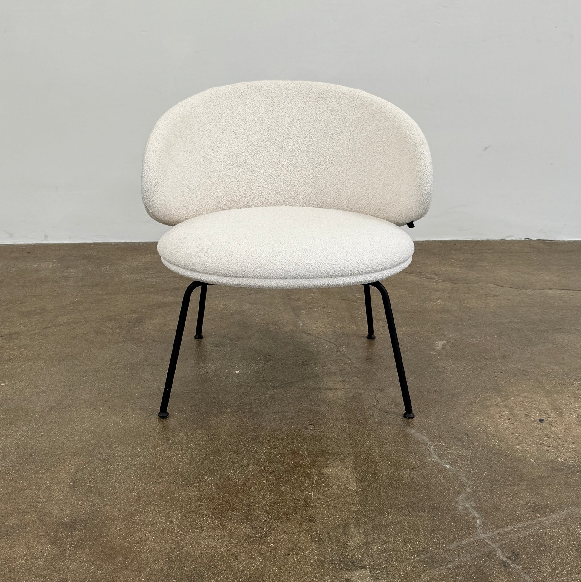 Two Arrmet Belle Lounge chairs by Arrmet, featuring white cushions, rounded backs, and black metal legs, are placed side by side on a brown concrete floor against a plain light gray wall.