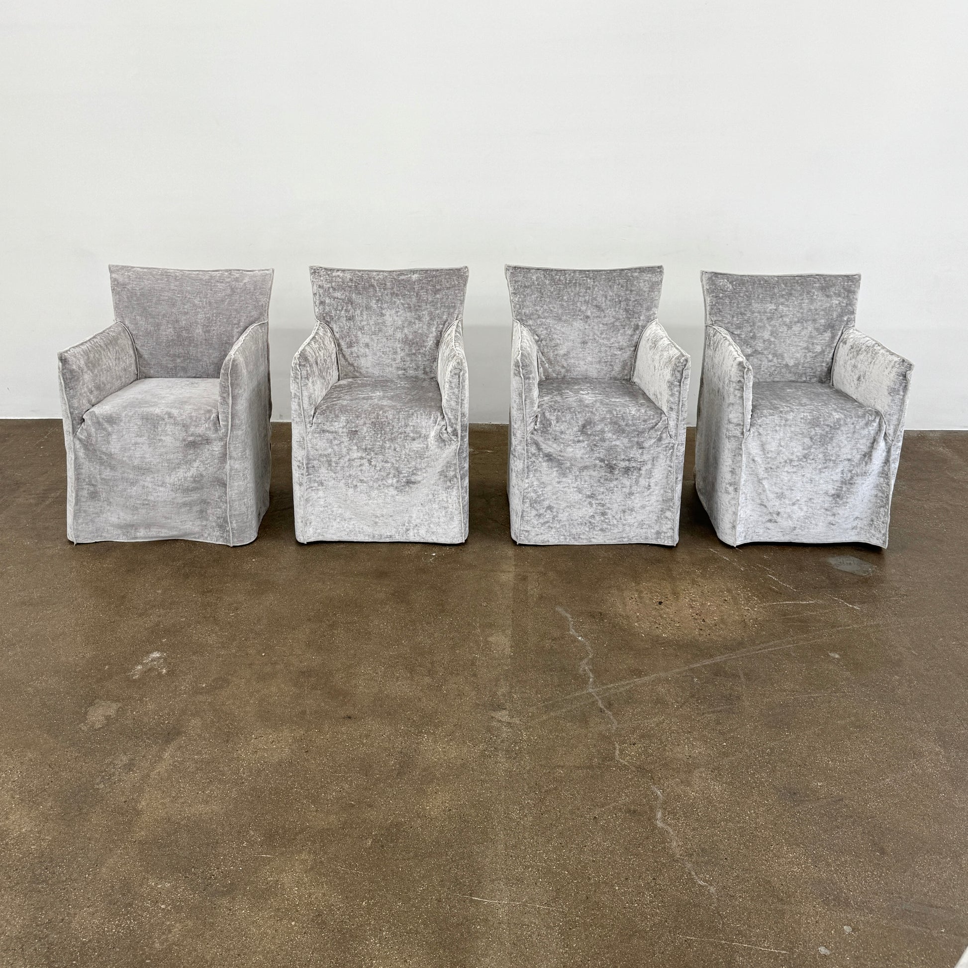 Four Gervasoni Ghost 25 Dining Armchairs by Gervasoni, featuring straight backs and skirted slipcovers in silver velvet, are arranged side by side against a white background for an elegant look.