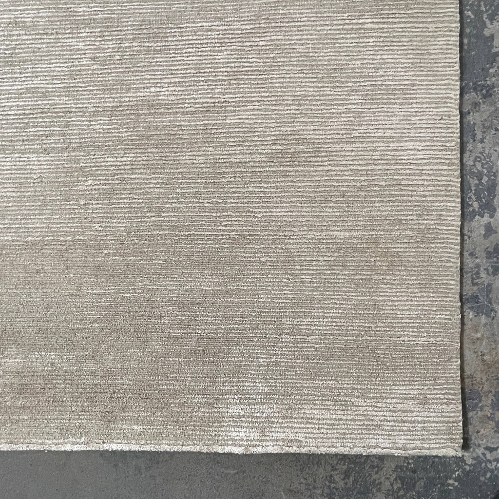 A close-up of a Delinear beige, hand-knotted textured carpet with a corner folded over, displayed against a white background.