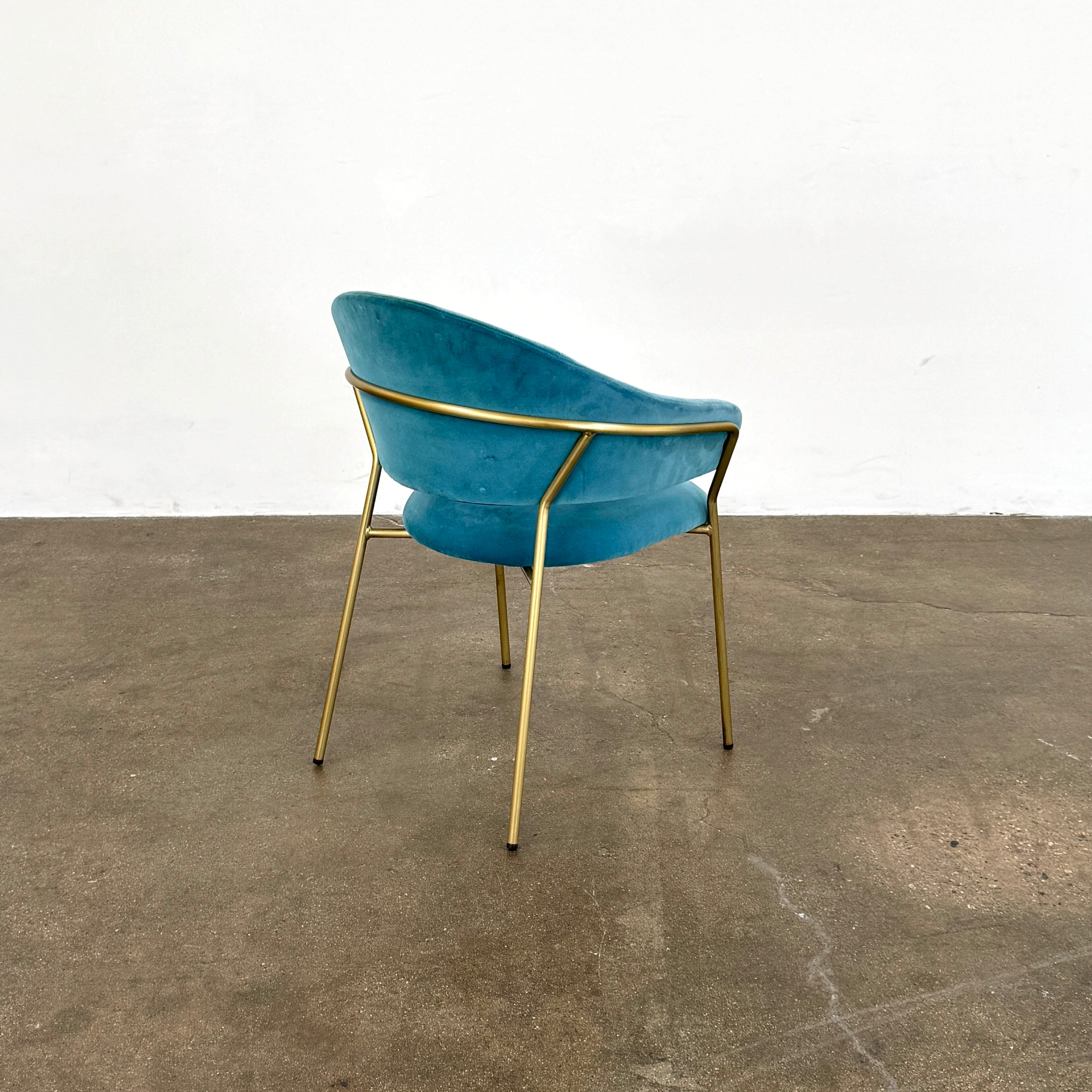 Two Pedrali Jazz Chairs by Pedrali, featuring customizable teal velvet upholstery and gold metal legs, sit side by side on a concrete floor against a white wall, highlighting their stylish steel structure.