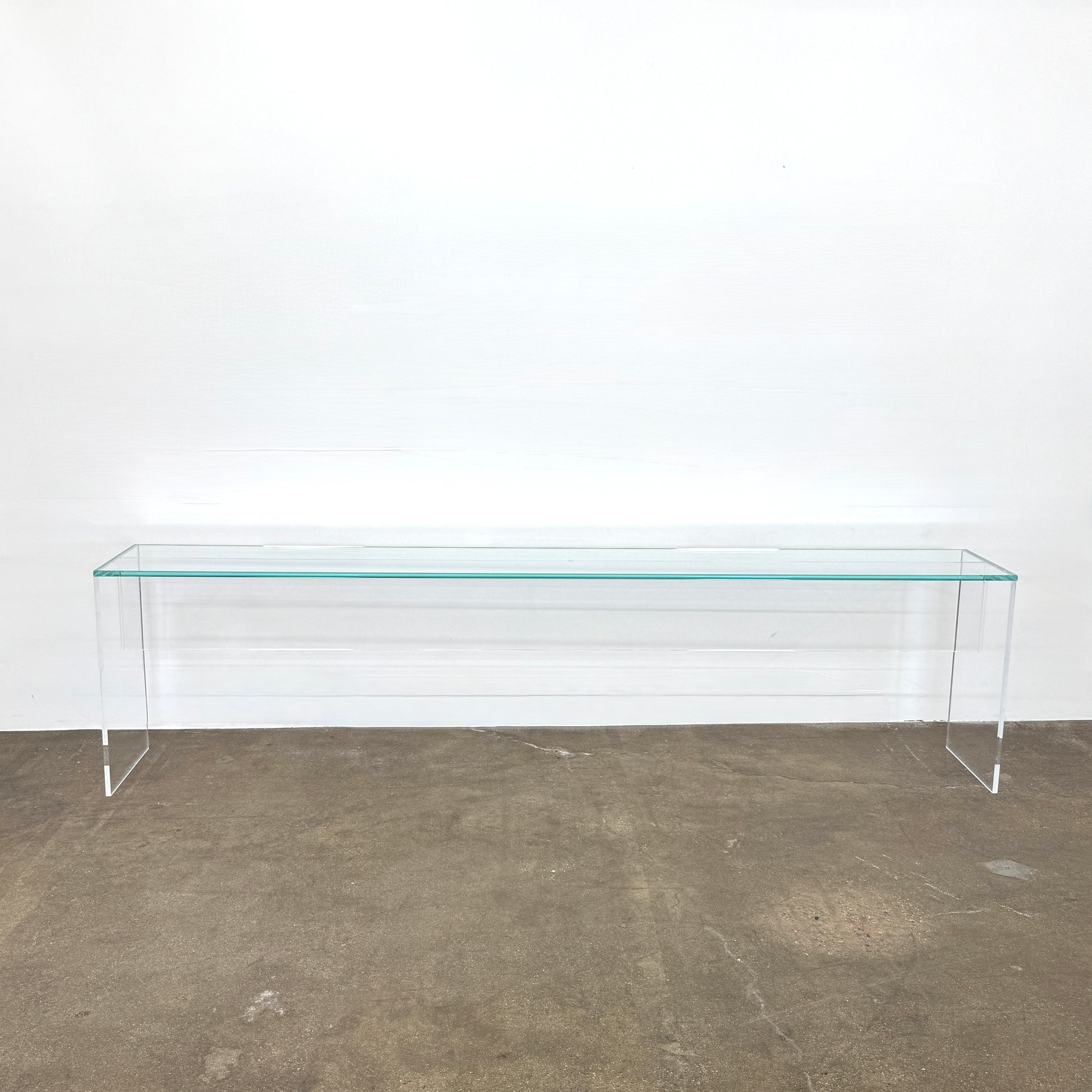 The Jacobson Vero Glass Console by Jacobson features clear rectangular glass surfaces and an acrylic base, elegantly displayed on a bare concrete floor against a plain white wall.
