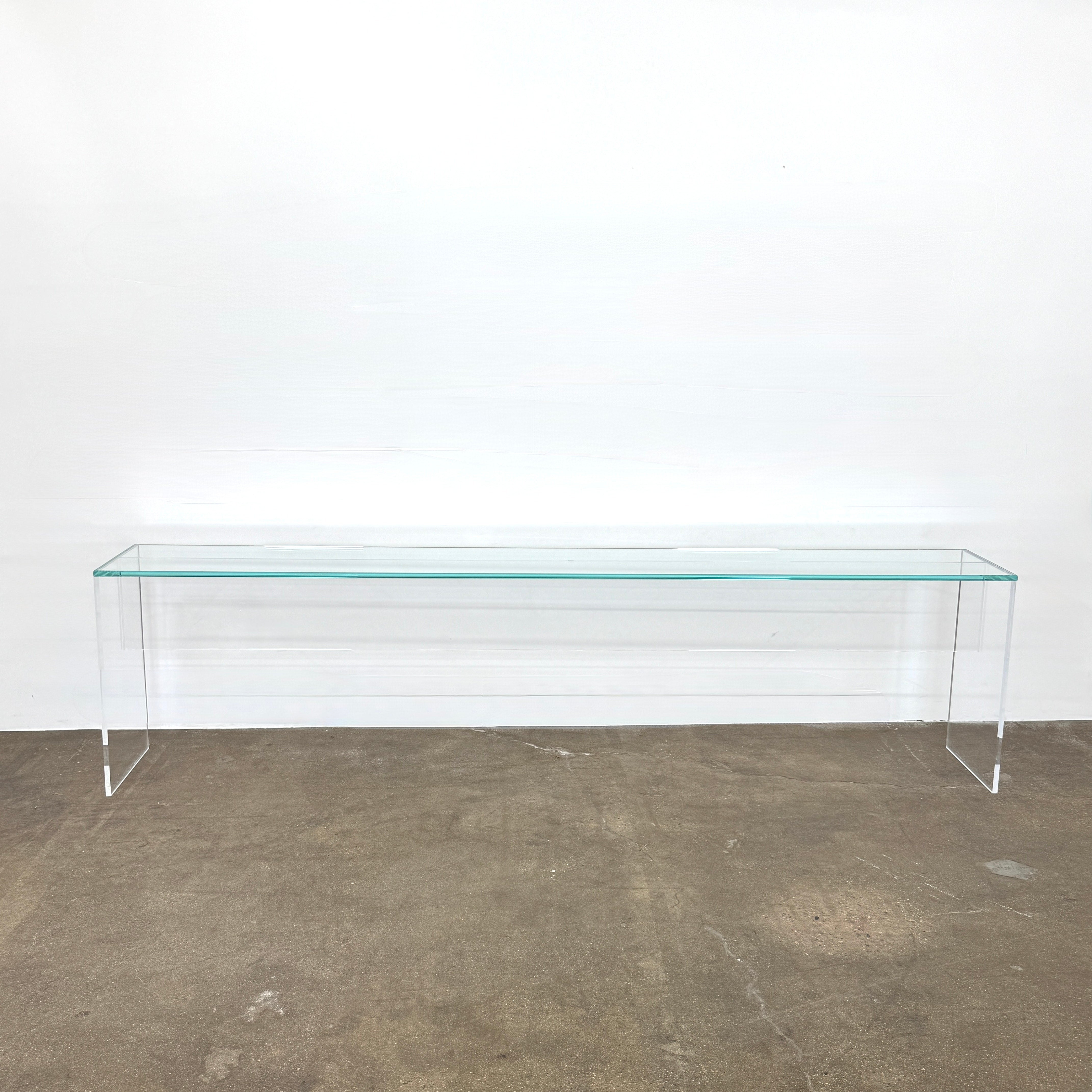 The Jacobson Vero Glass Console by Jacobson features clear rectangular glass surfaces and an acrylic base, elegantly displayed on a bare concrete floor against a plain white wall.