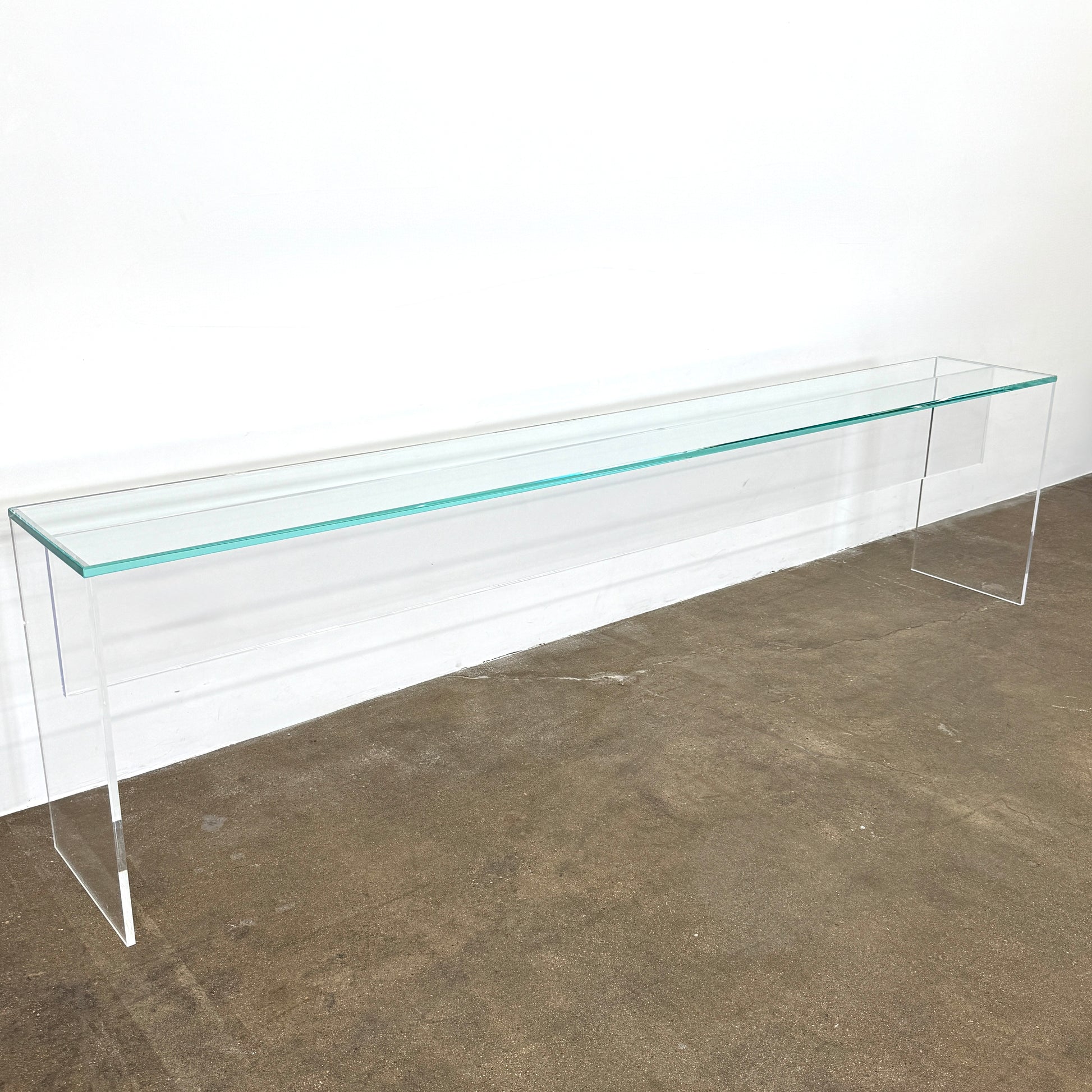 The Jacobson Vero Glass Console by Jacobson features clear rectangular glass surfaces and an acrylic base, elegantly displayed on a bare concrete floor against a plain white wall.