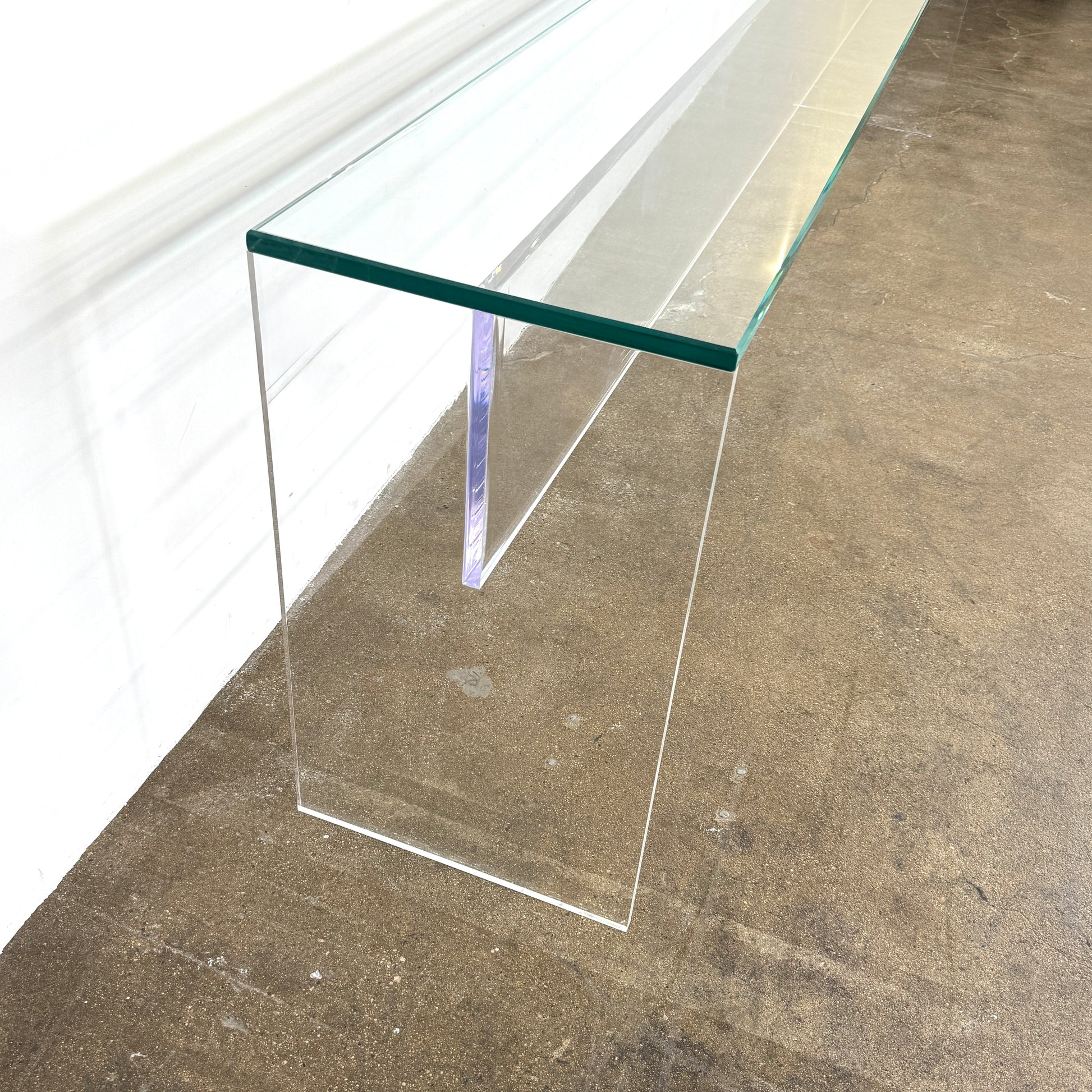 The Jacobson Vero Glass Console by Jacobson features clear rectangular glass surfaces and an acrylic base, elegantly displayed on a bare concrete floor against a plain white wall.