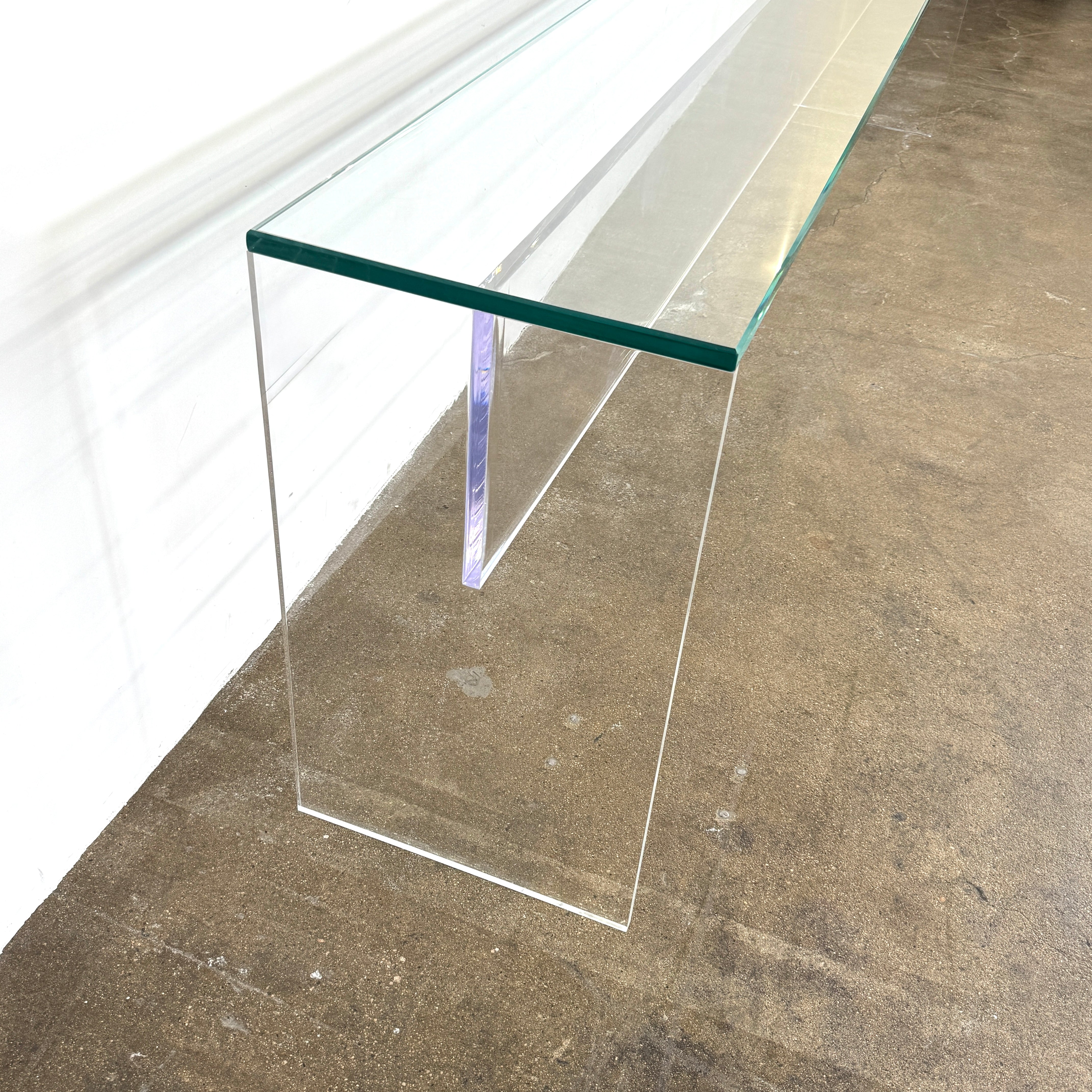 The Jacobson Vero Glass Console by Jacobson features clear rectangular glass surfaces and an acrylic base, elegantly displayed on a bare concrete floor against a plain white wall.