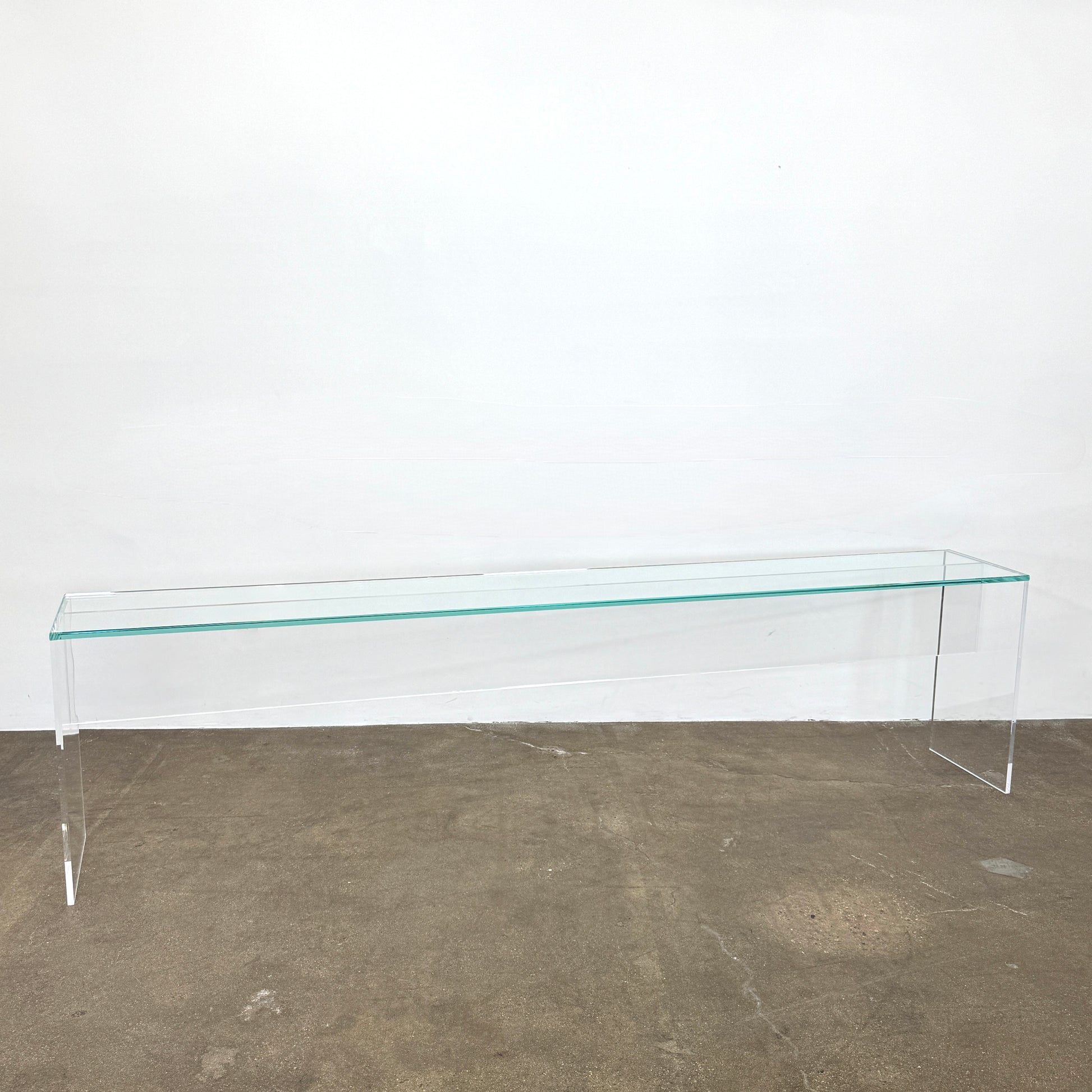 The Jacobson Vero Glass Console by Jacobson features clear rectangular glass surfaces and an acrylic base, elegantly displayed on a bare concrete floor against a plain white wall.