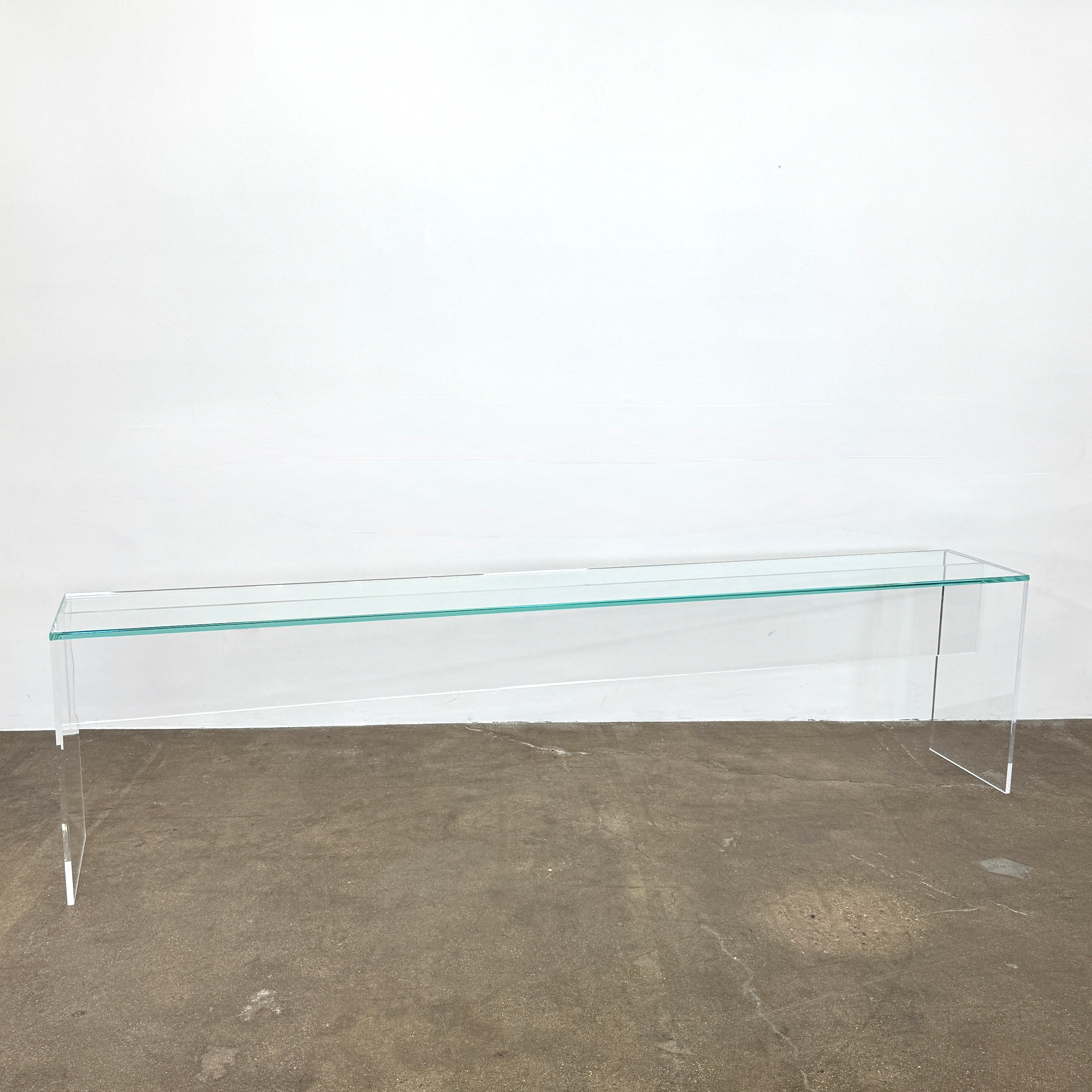 The Jacobson Vero Glass Console by Jacobson features clear rectangular glass surfaces and an acrylic base, elegantly displayed on a bare concrete floor against a plain white wall.