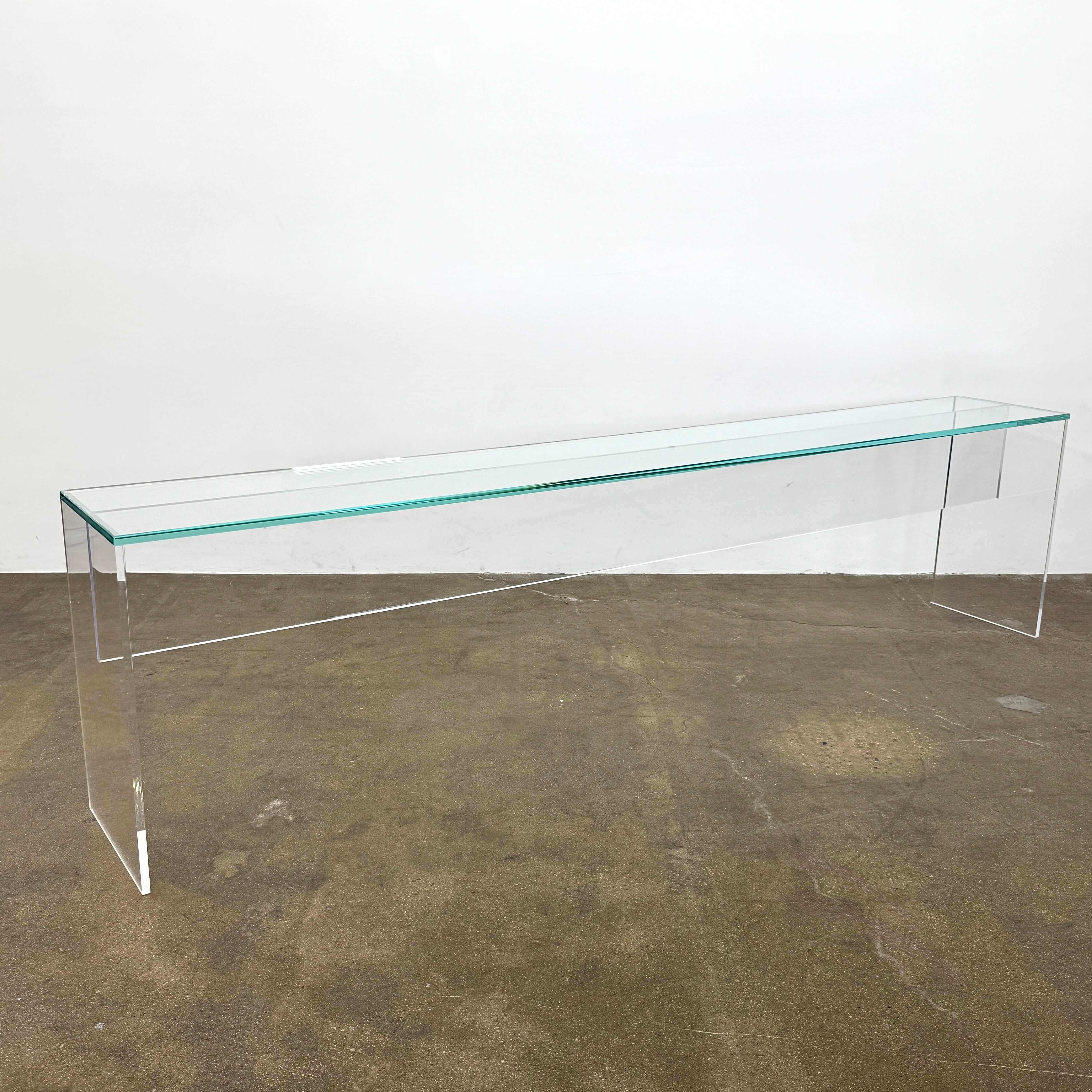 The Jacobson Vero Glass Console by Jacobson features clear rectangular glass surfaces and an acrylic base, elegantly displayed on a bare concrete floor against a plain white wall.