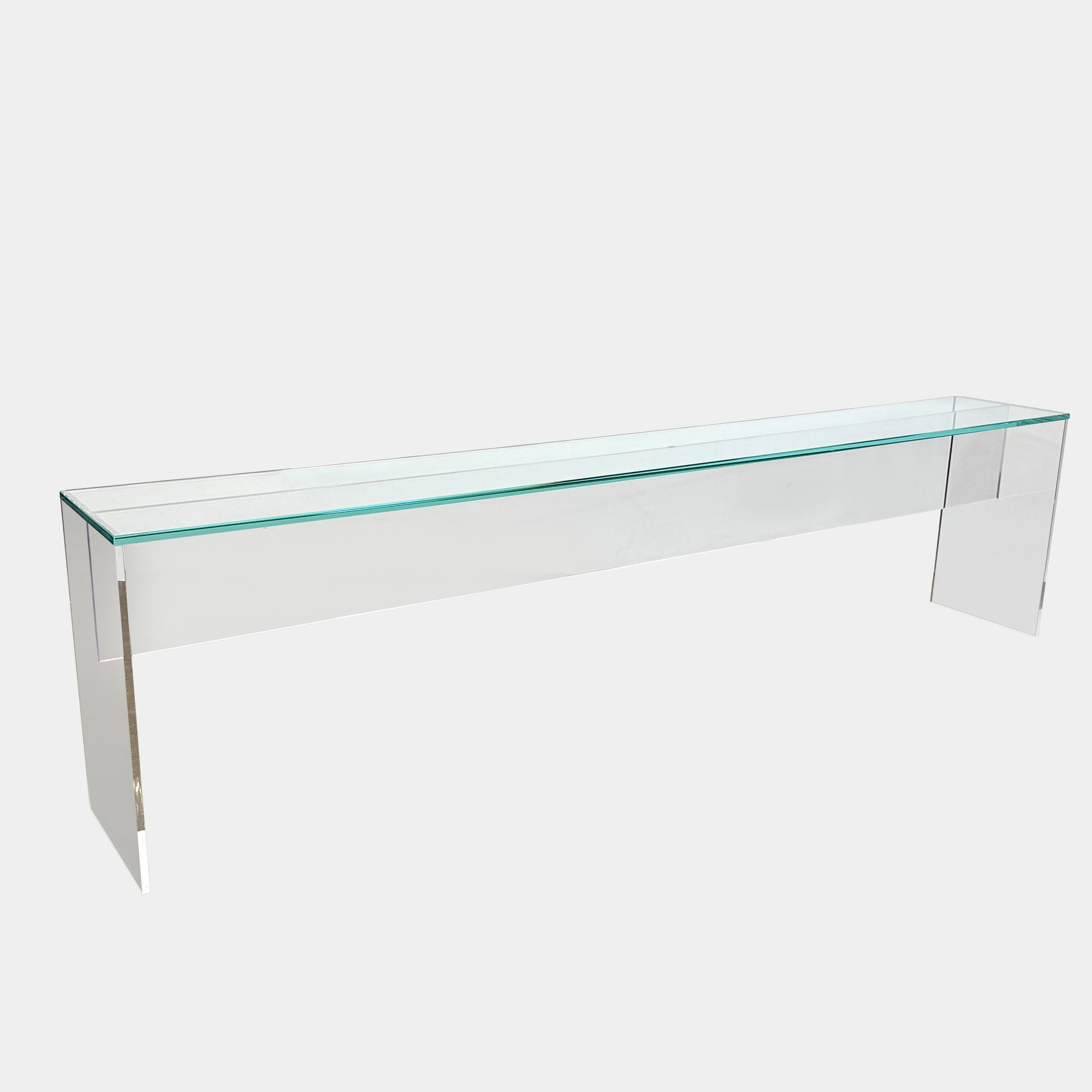 The Jacobson Vero glass console 118” from Modern Resale features a clear, minimalist design and is shown placed on a concrete floor against a white wall.