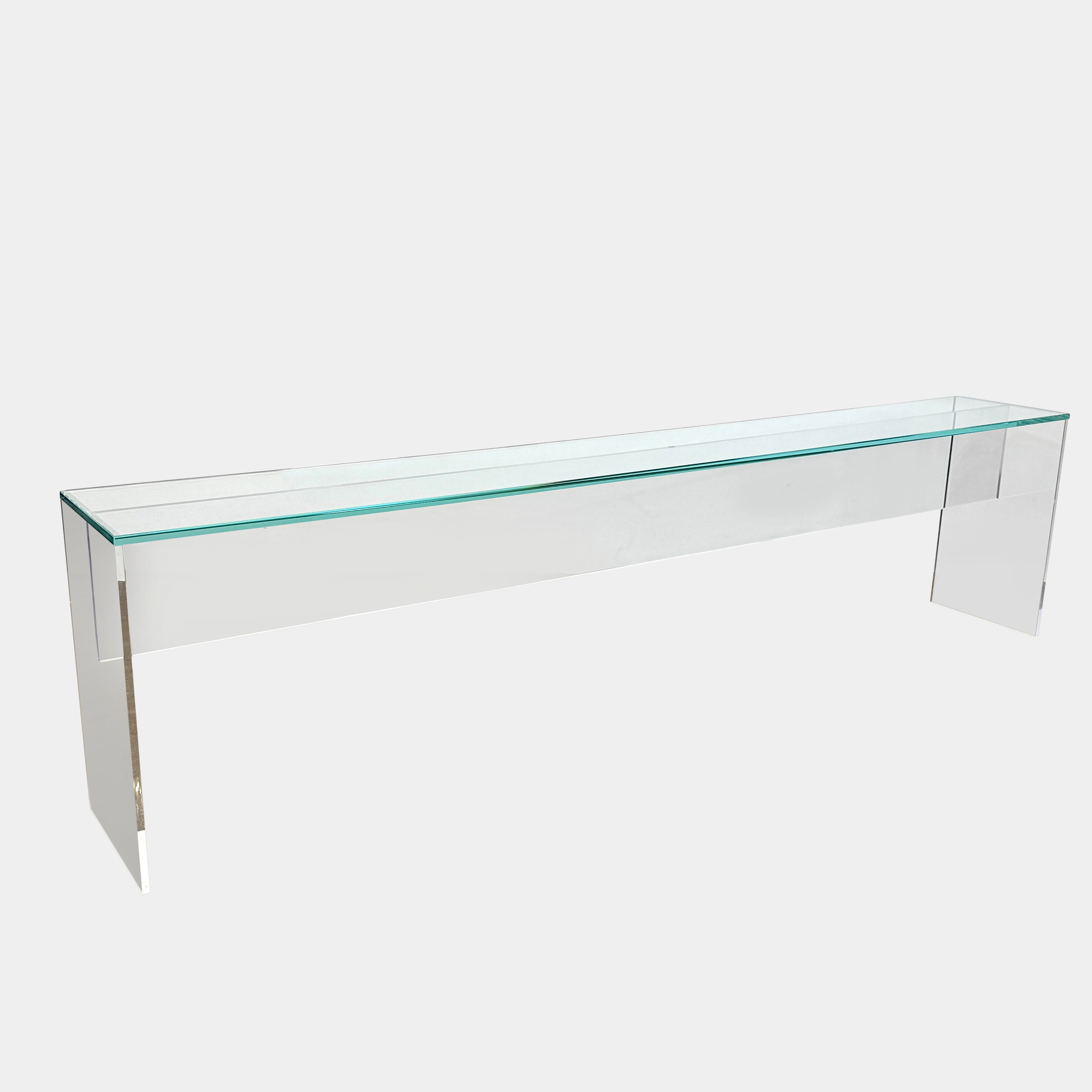 The Jacobson Vero glass console 118” from Modern Resale features a clear, minimalist design and is shown placed on a concrete floor against a white wall.