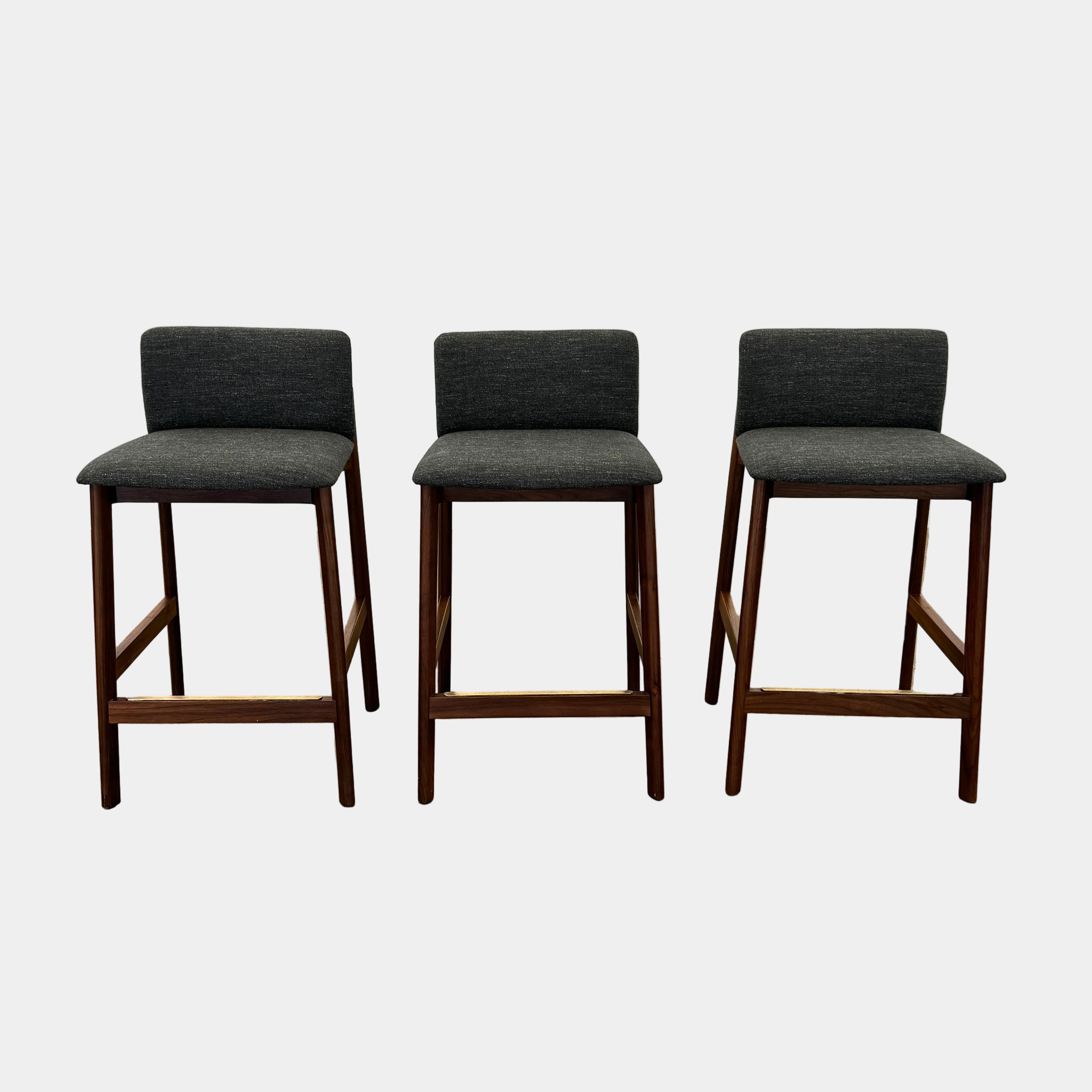 Three DWR Contour Counter Stools (Set of 3) with dark grey upholstered seats and walnut frames are arranged in a row on a polished concrete floor against a plain white wall.