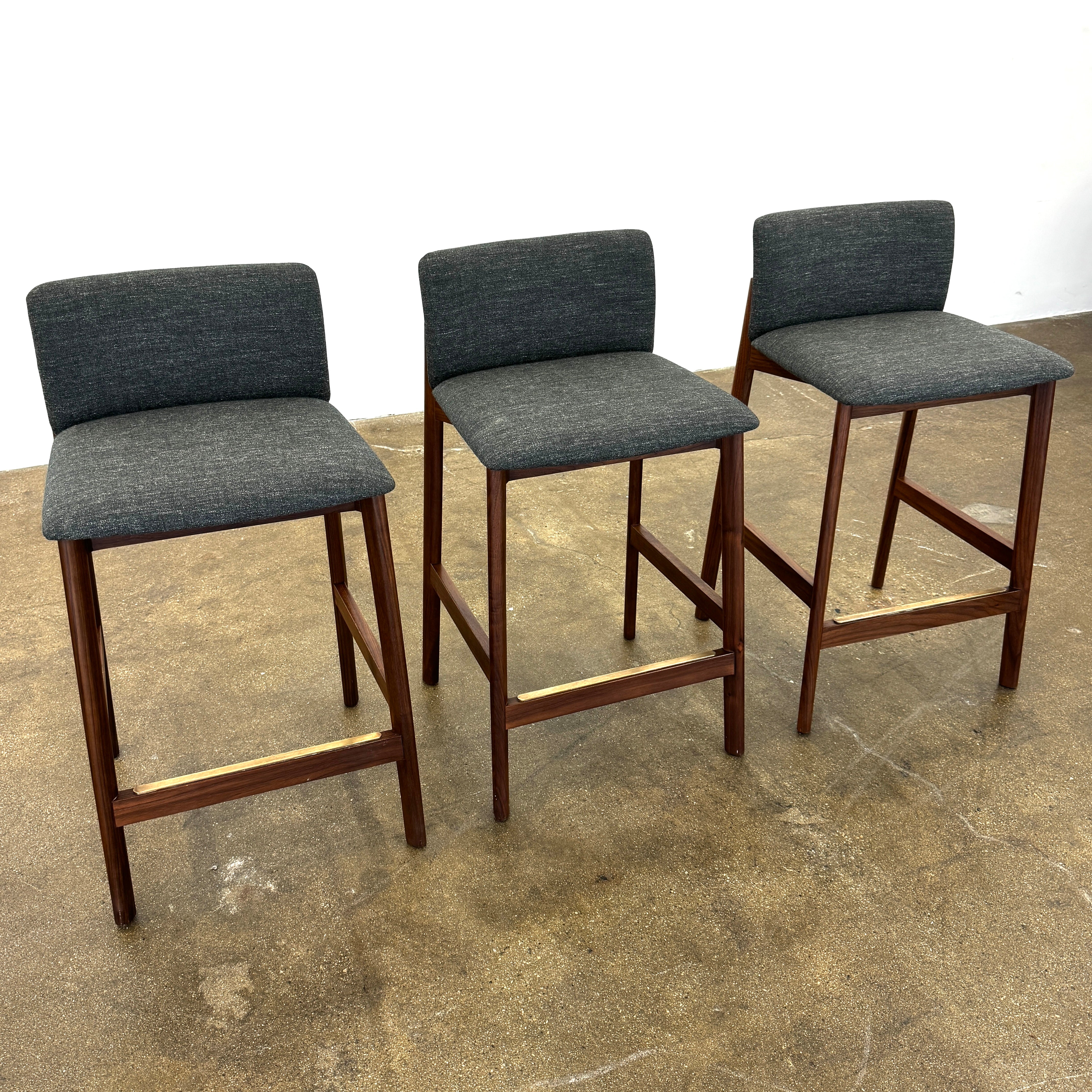 Three DWR Contour Counter Stools (Set of 3) with dark grey upholstered seats and walnut frames are arranged in a row on a polished concrete floor against a plain white wall.