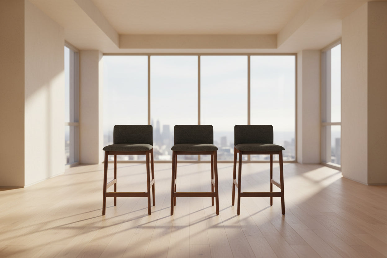 Three DWR Contour Counter Stools (Set of 3) with dark grey upholstered seats and walnut frames are arranged in a row on a polished concrete floor against a plain white wall.
