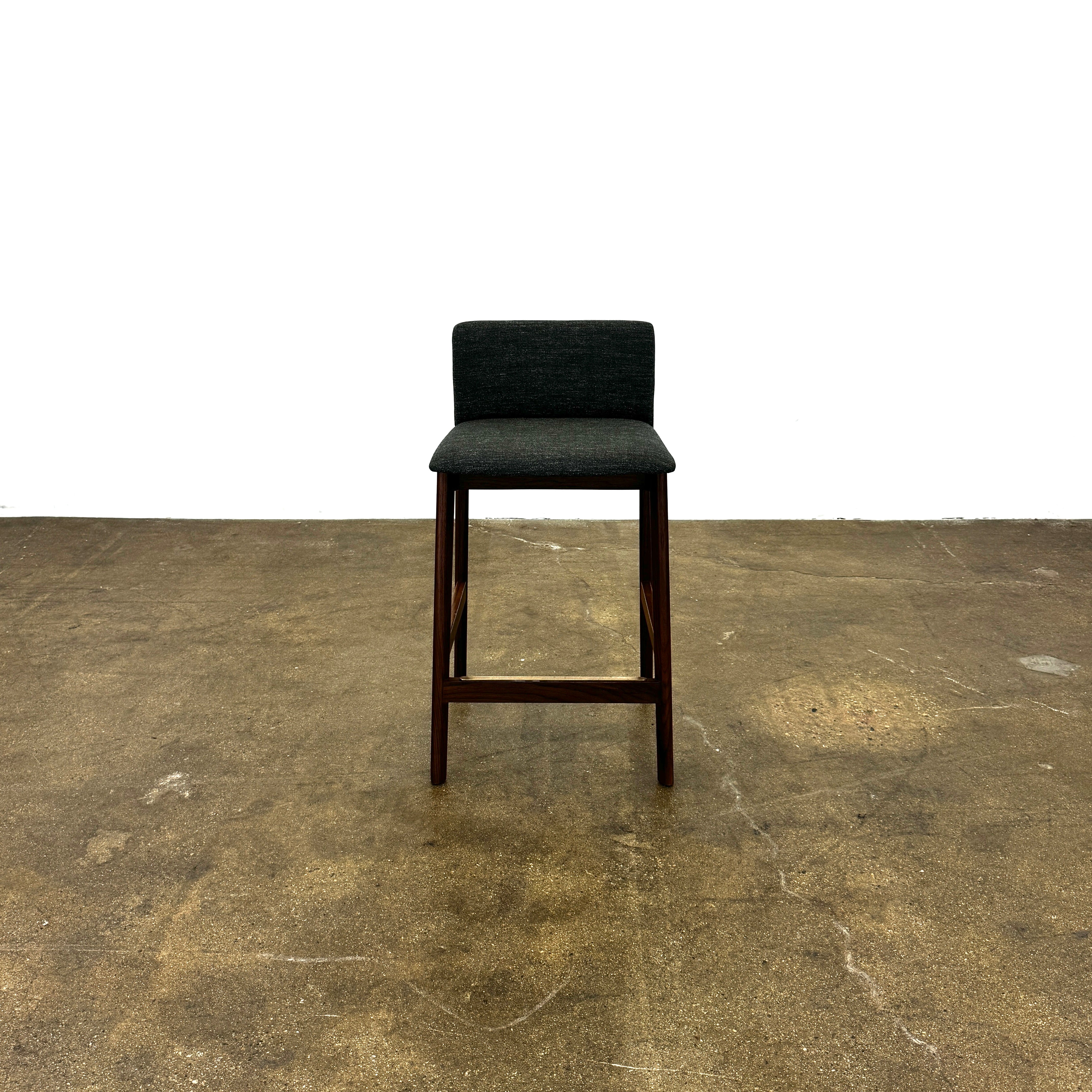 Three DWR Contour Counter Stools (Set of 3) with dark grey upholstered seats and walnut frames are arranged in a row on a polished concrete floor against a plain white wall.