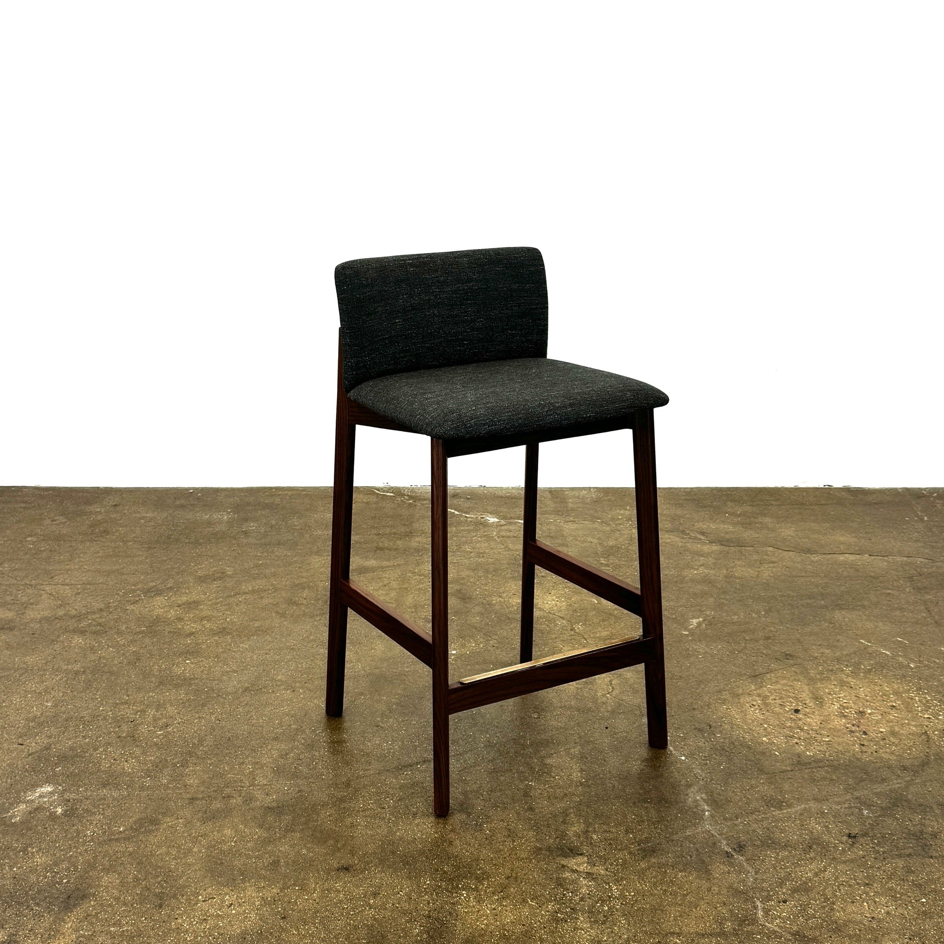 Three DWR Contour Counter Stools (Set of 3) with dark grey upholstered seats and walnut frames are arranged in a row on a polished concrete floor against a plain white wall.