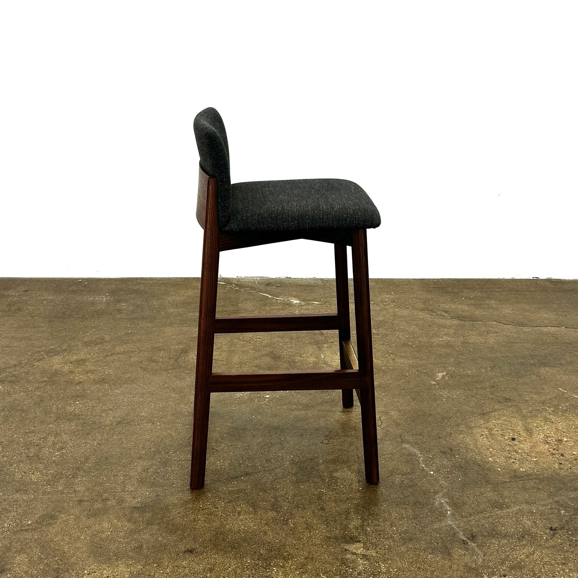 Three DWR Contour Counter Stools (Set of 3) with dark grey upholstered seats and walnut frames are arranged in a row on a polished concrete floor against a plain white wall.