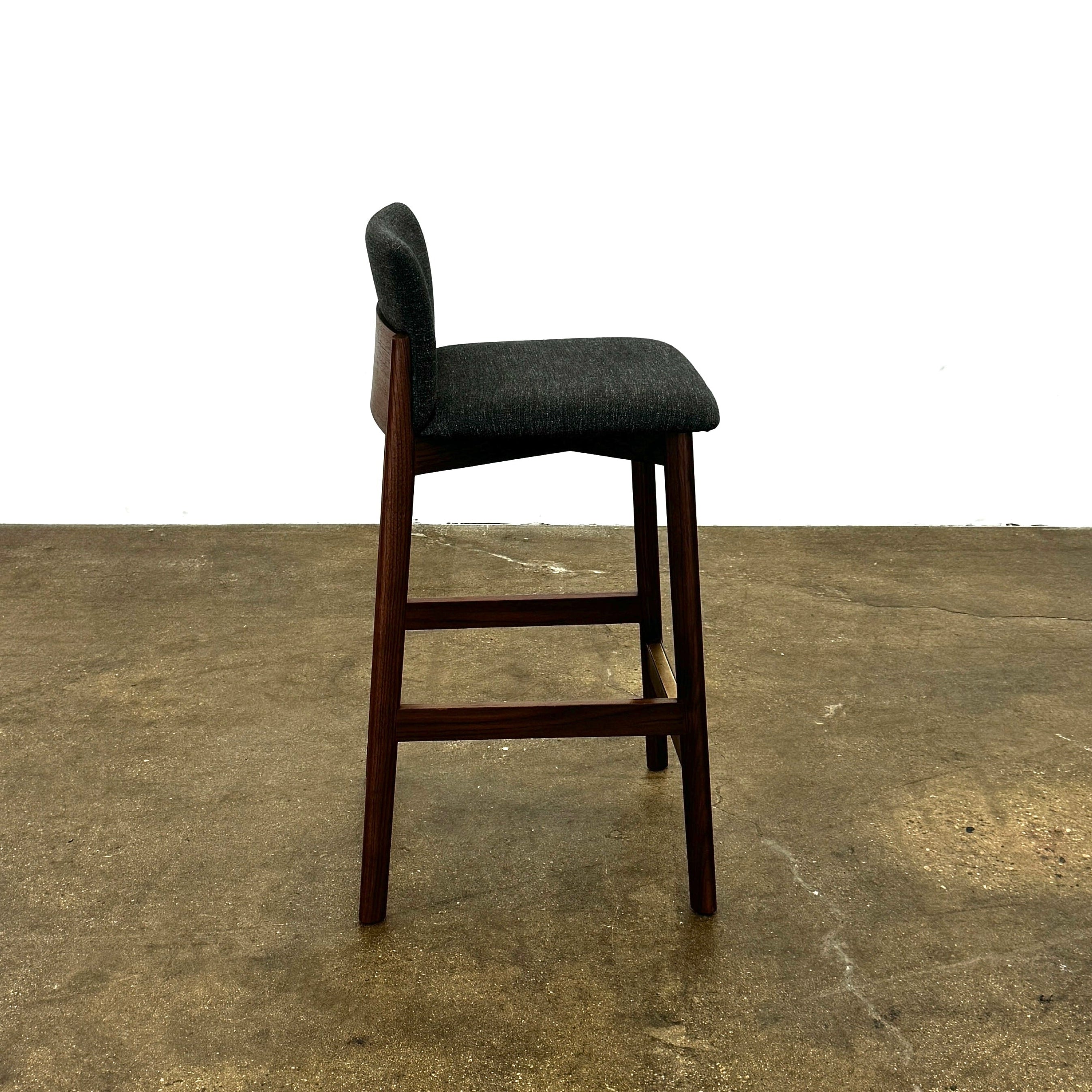 Three DWR Contour Counter Stools (Set of 3) with dark grey upholstered seats and walnut frames are arranged in a row on a polished concrete floor against a plain white wall.