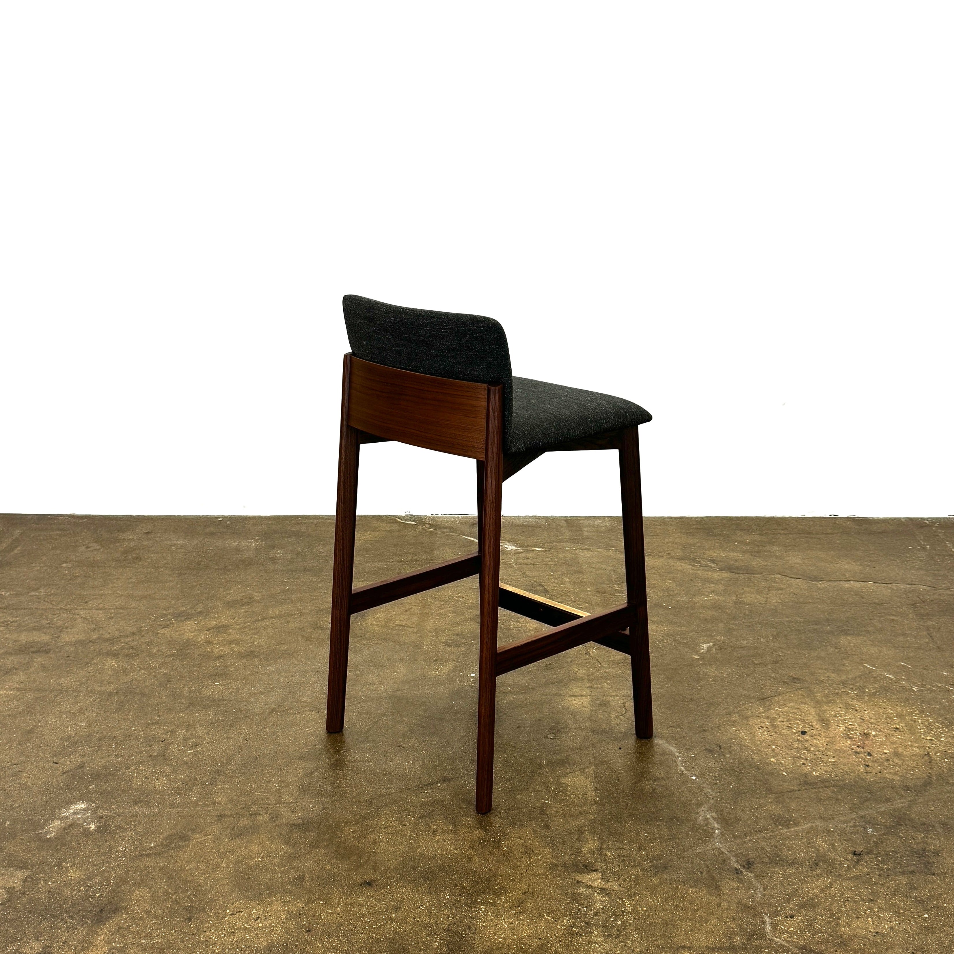 Three DWR Contour Counter Stools (Set of 3) with dark grey upholstered seats and walnut frames are arranged in a row on a polished concrete floor against a plain white wall.
