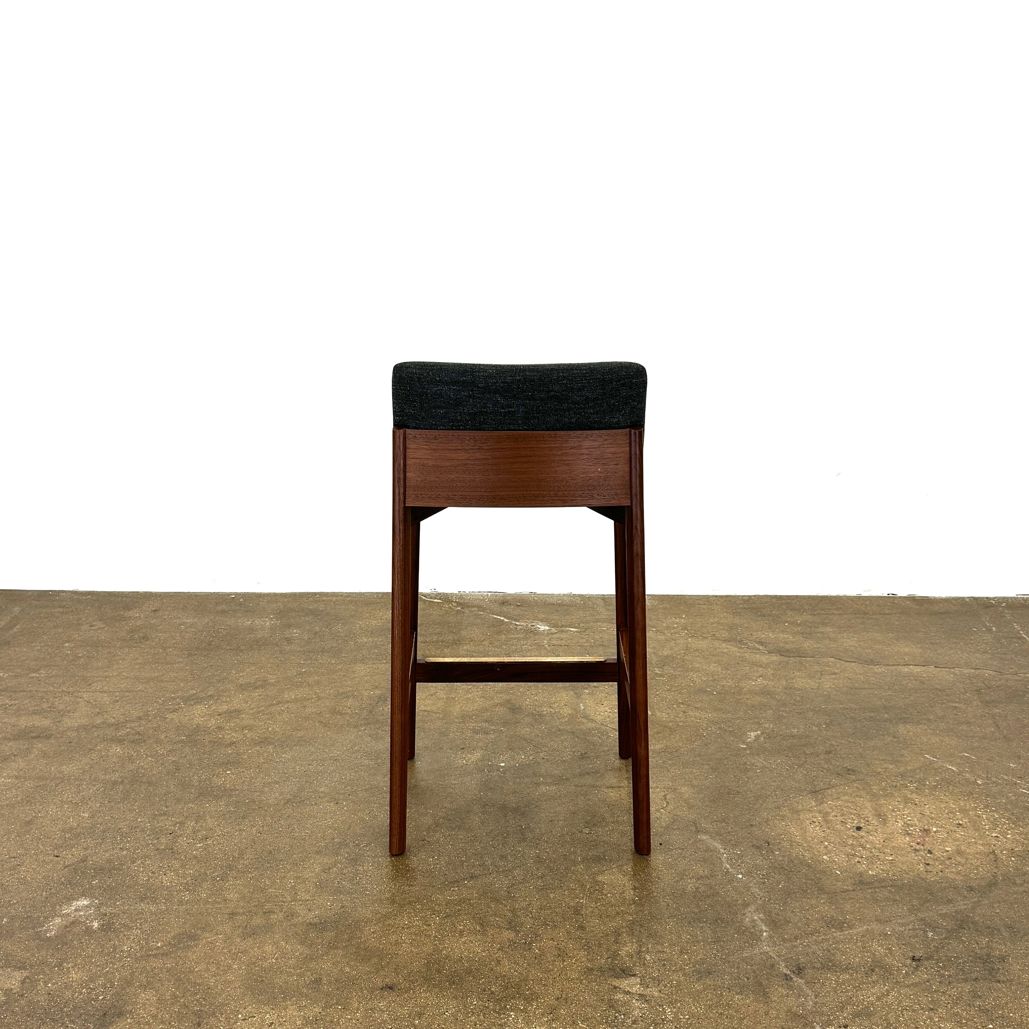 Three DWR Contour Counter Stools (Set of 3) with dark grey upholstered seats and walnut frames are arranged in a row on a polished concrete floor against a plain white wall.