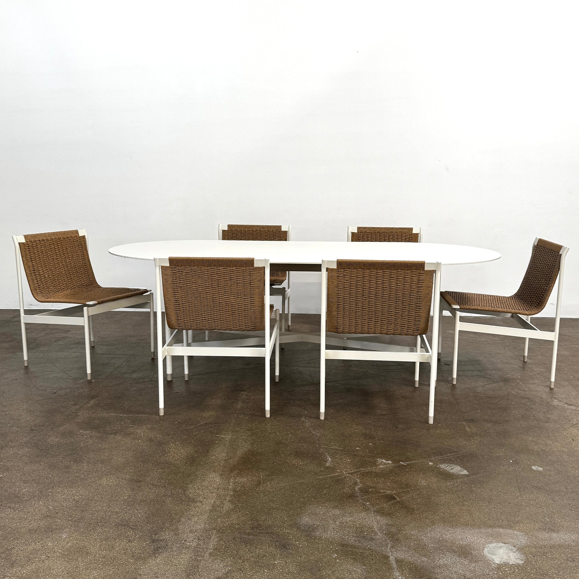 The MillerKnoll Outdoor Sommer Oval Dining Table for DWR, paired with six brown wicker chairs on a polished concrete floor against a white wall, is ideal for fans of modern patio furniture and minimalist design.