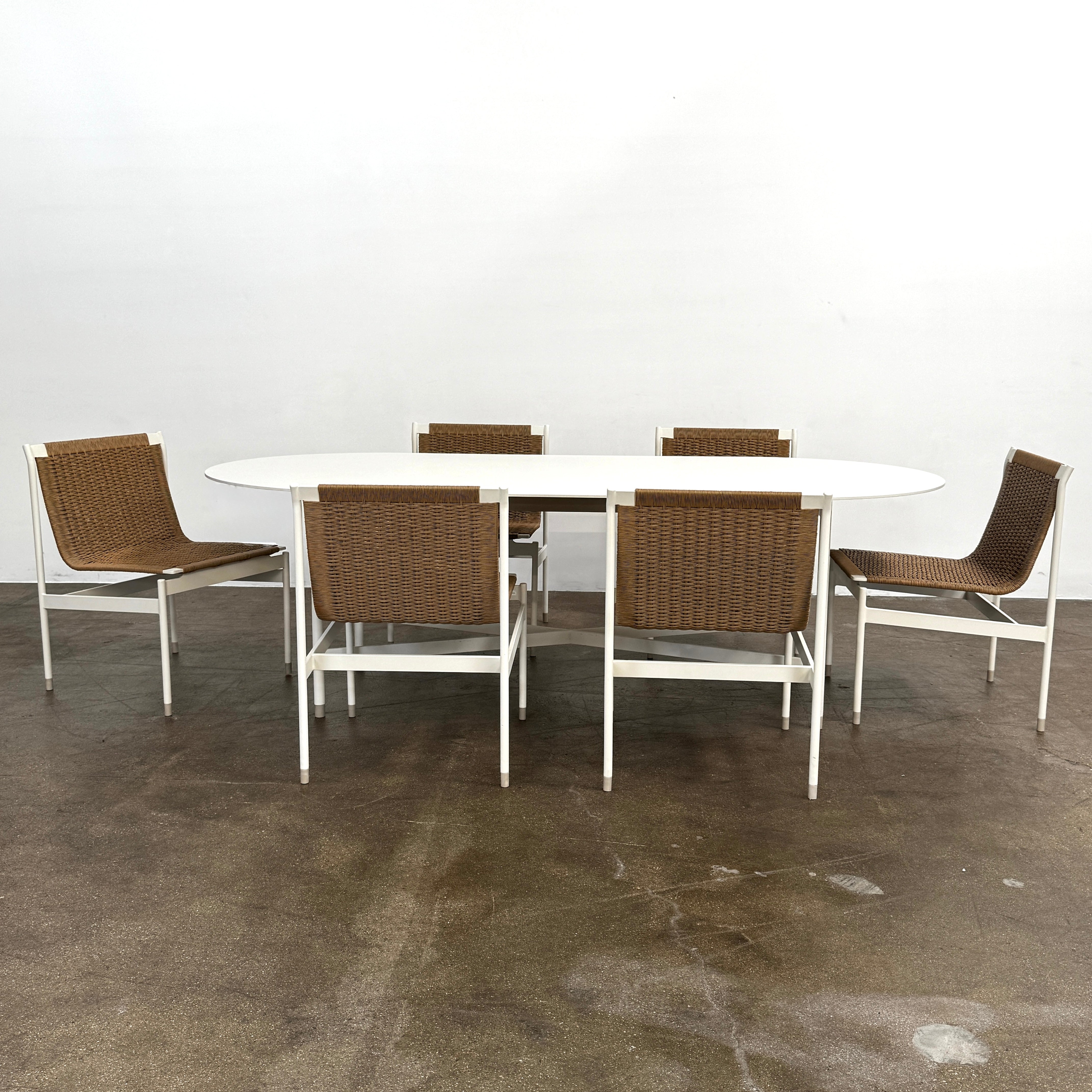 The MillerKnoll Outdoor Sommer Oval Dining Table for DWR, paired with six brown wicker chairs on a polished concrete floor against a white wall, is ideal for fans of modern patio furniture and minimalist design.
