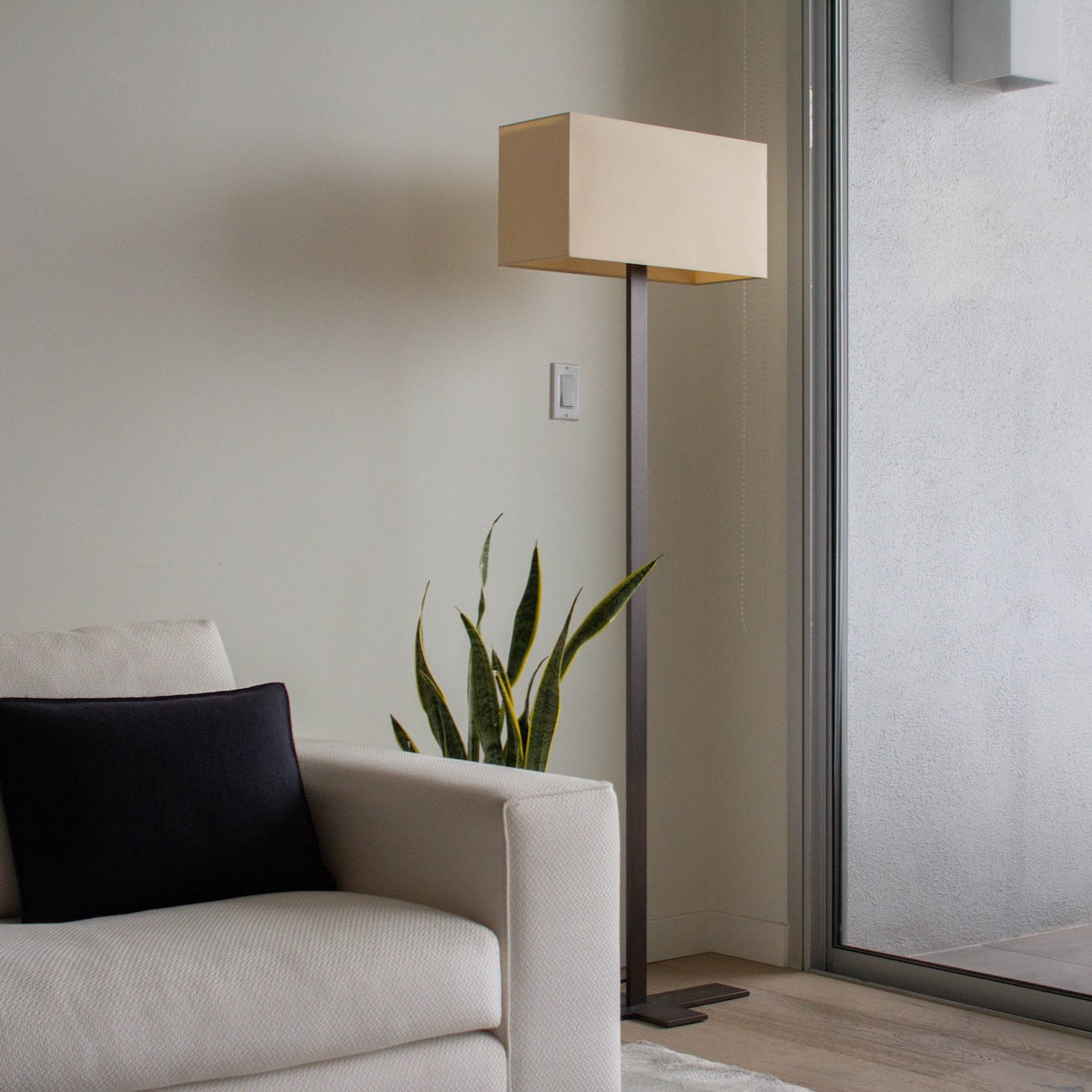 The Maxalto Leukon Tall Floor Lamp by Maxalto, featuring a minimalist design with a rectangular beige shade and a bronze nickel stand, stands on a bare concrete floor, plugged into an electrical outlet and casting ambient light throughout the space.