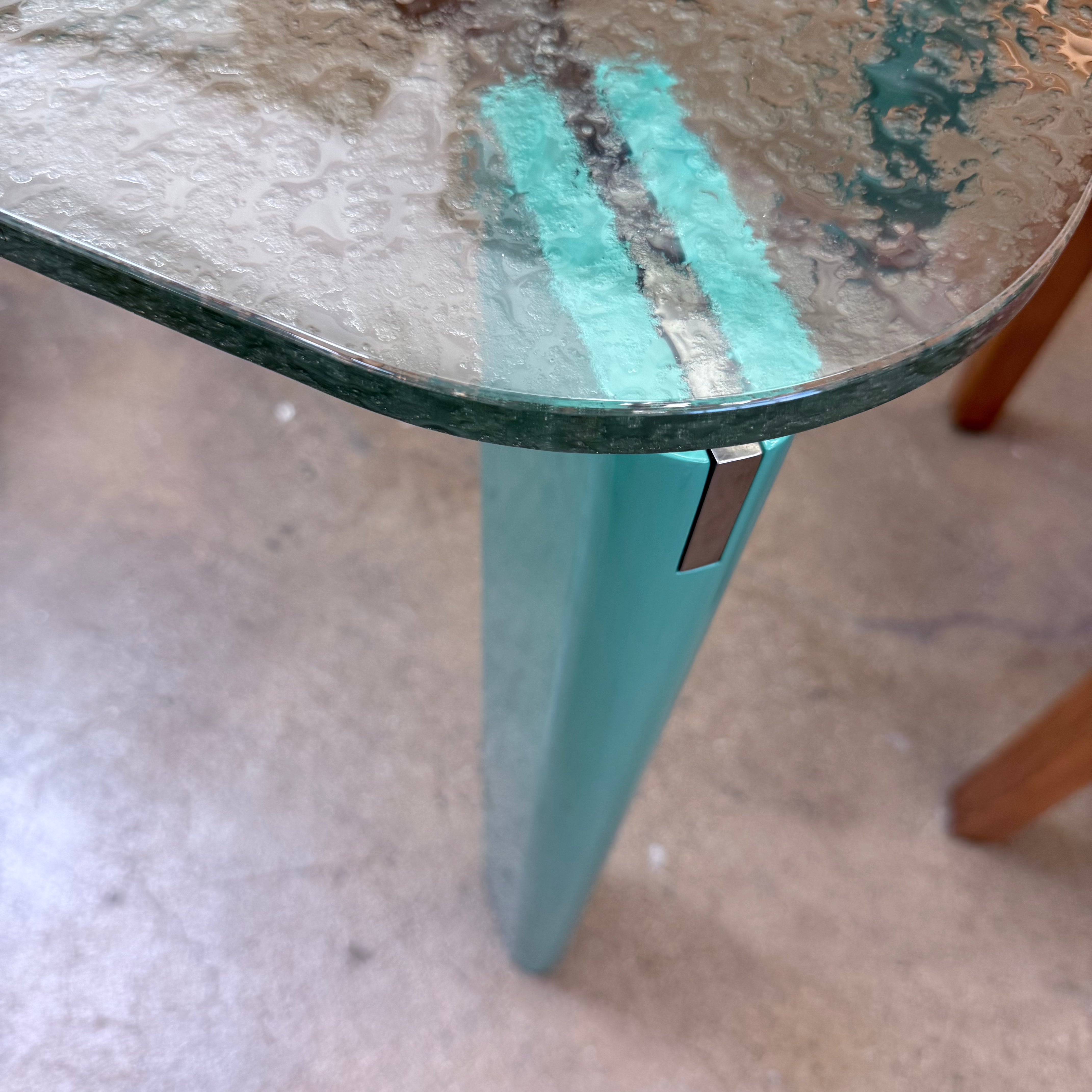 The B&B Italia Oskar Dining Table by B&B Italia features a rectangular glass top with four teal legs, displayed against a plain white background.