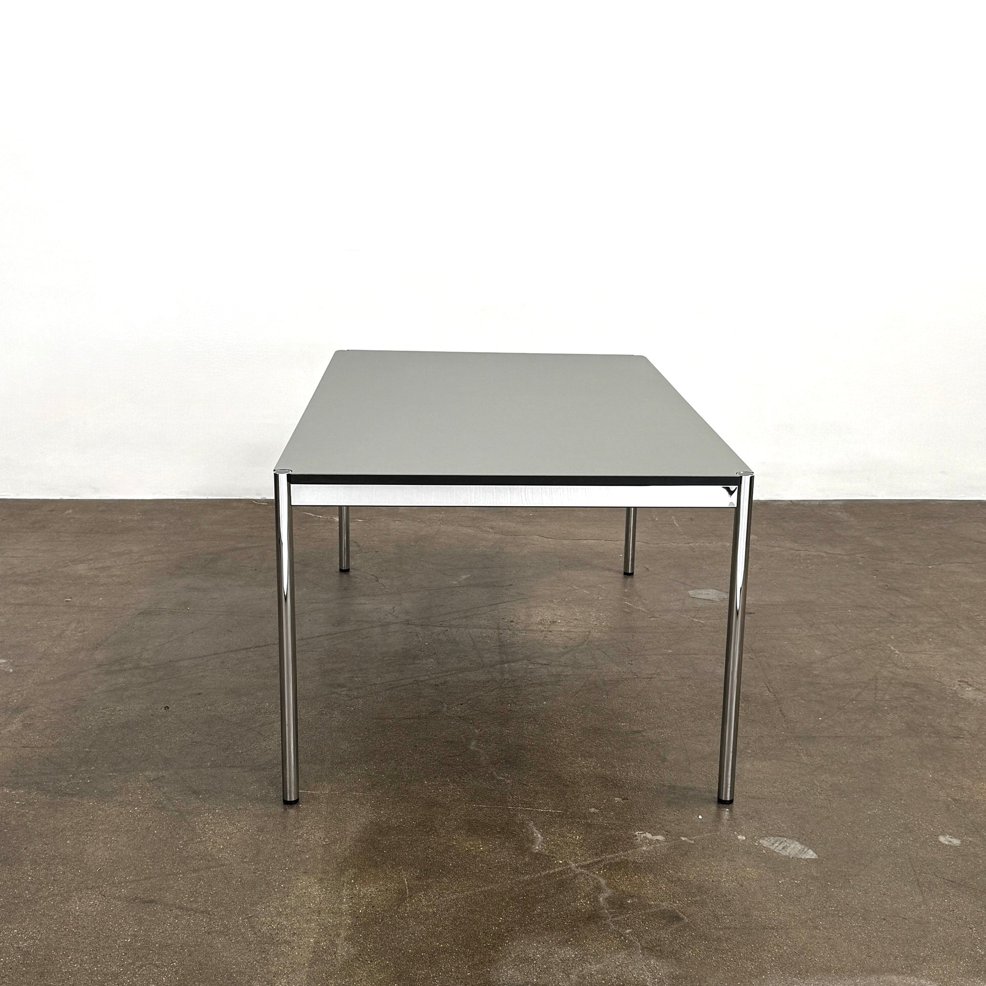 A modern mesh-back office chair sits at a USM Haller Work Table by USM Haller, which has a minimalist design and metal legs, placed on a concrete floor with a white background.