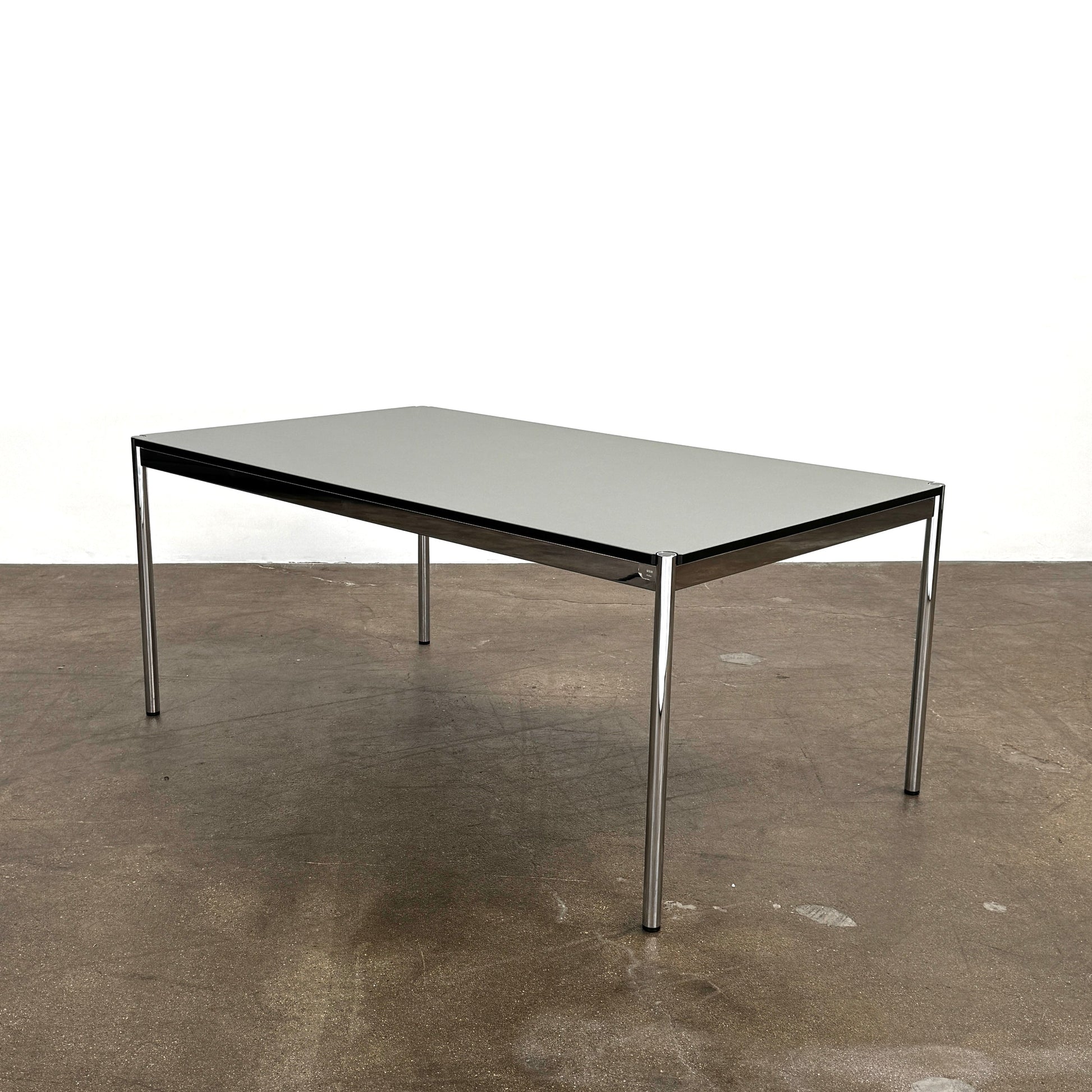 A modern mesh-back office chair sits at a USM Haller Work Table by USM Haller, which has a minimalist design and metal legs, placed on a concrete floor with a white background.